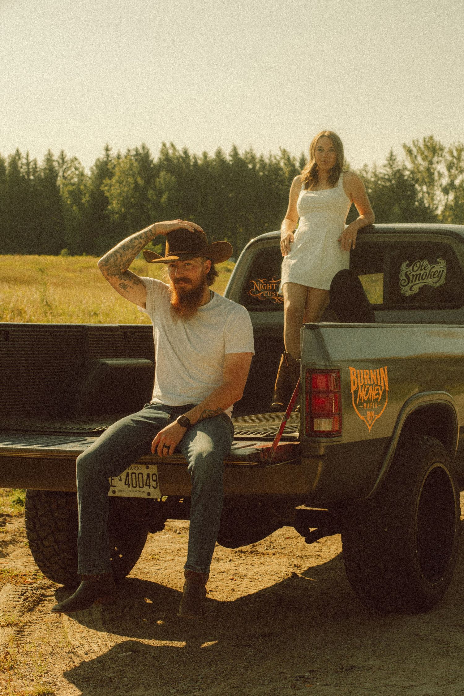 Bearded man in white tee sits on pickup truck tailgate while woman in white dress stands in truck bed, rural field backdrop.