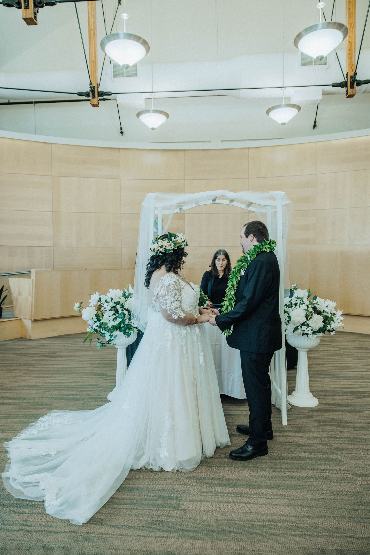 A bride and groom sharing their first dance in an elegant ballroom with wood flooring and white floral arrangements.