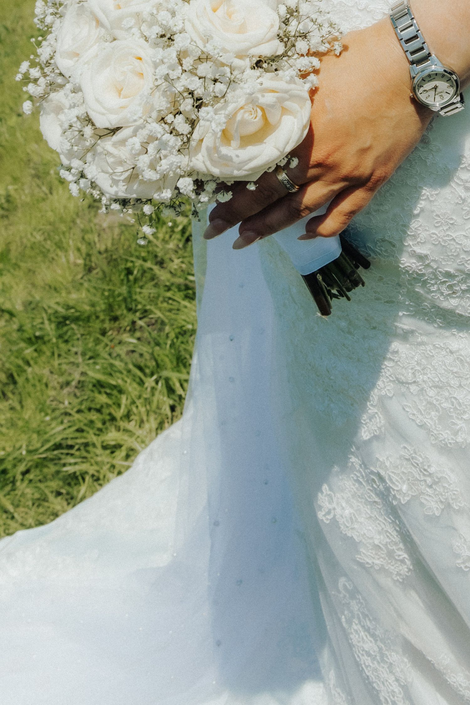Bride in white lace wedding dress holding elegant bouquet of white roses and baby's breath, wearing a silver watch.