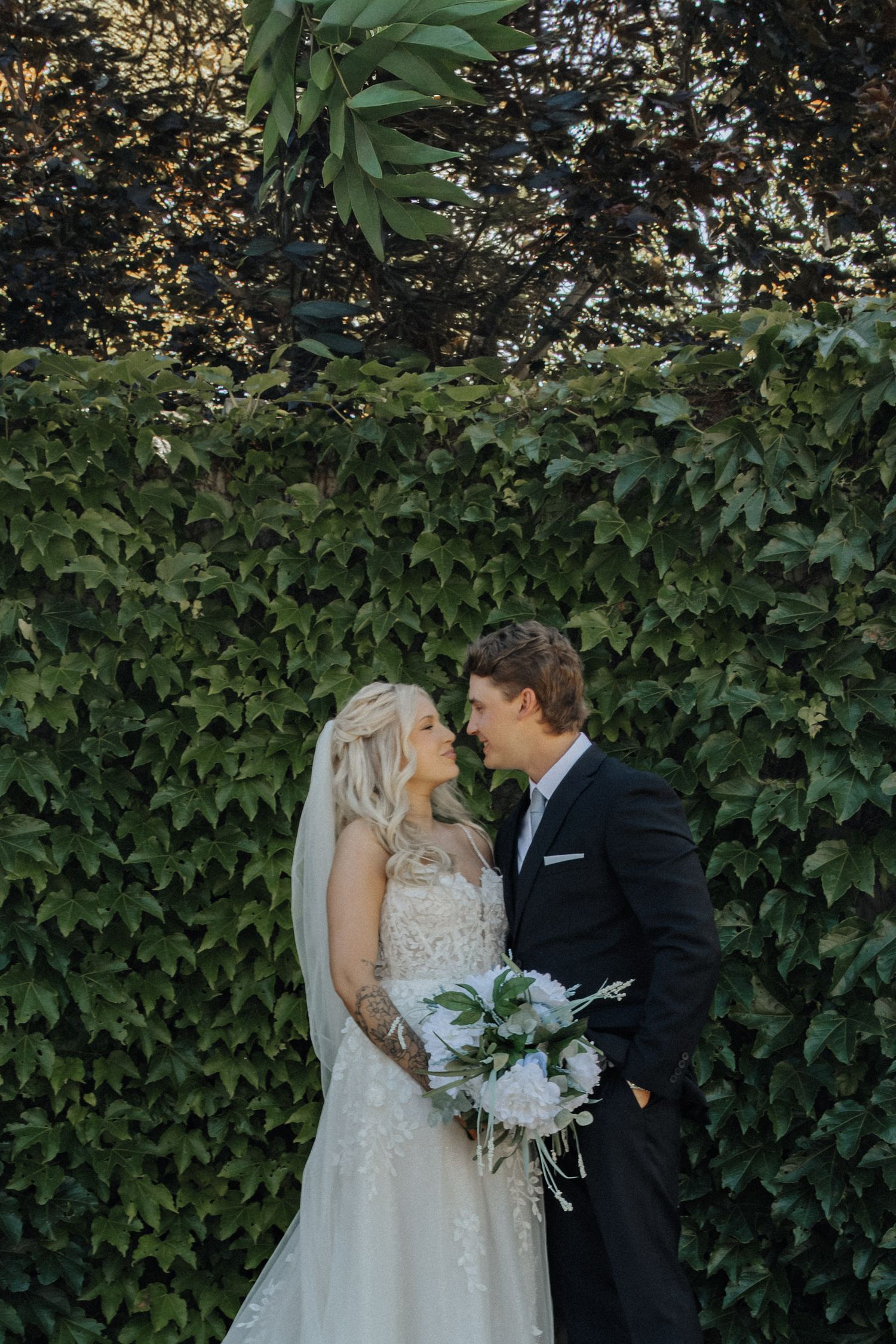 Bride and groom share a romantic moment in front of a lush green ivy wall, bride holding white bouquet.