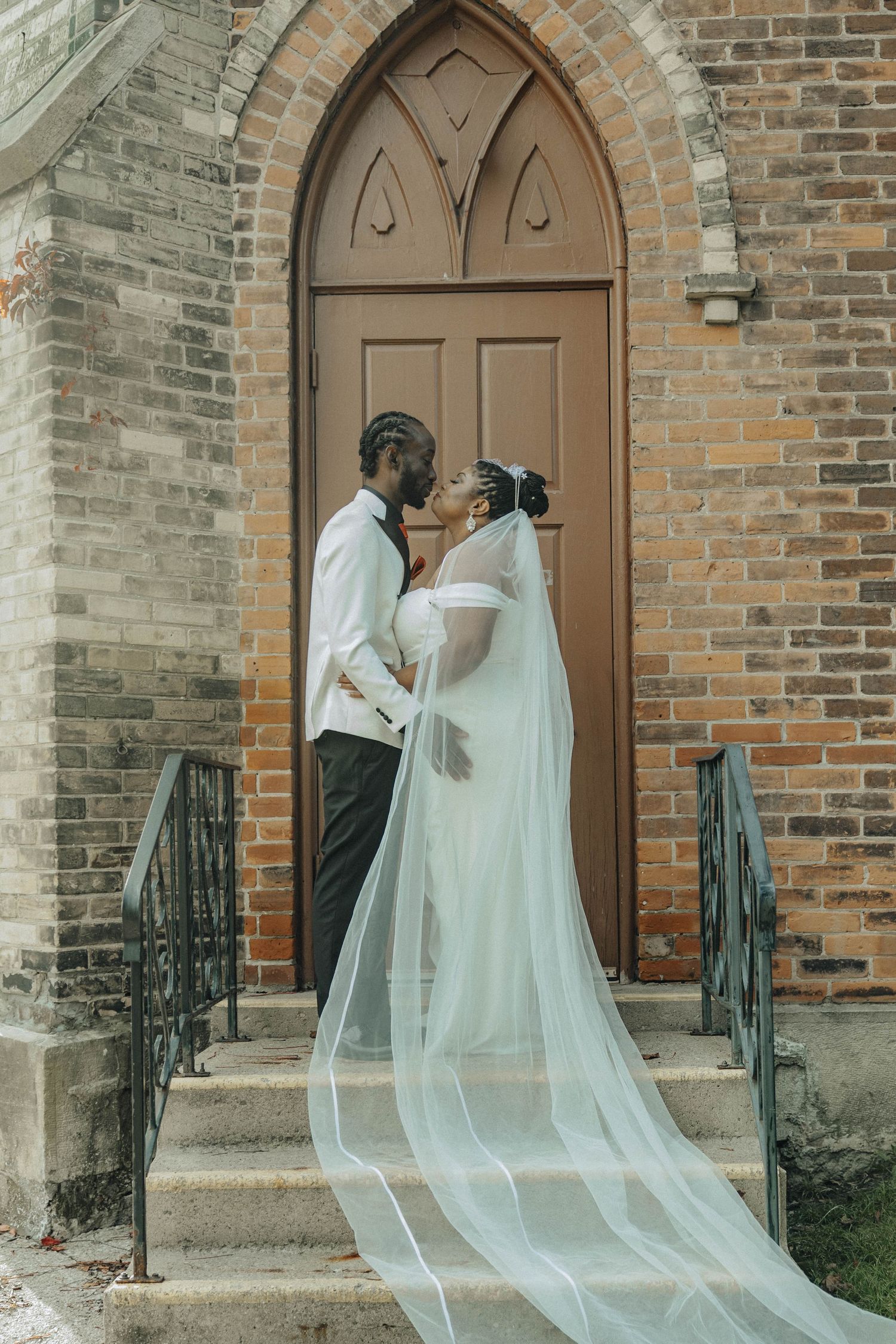 A flowing white bridal veil cascades down church steps against a rustic brick wall with arched wooden doors.