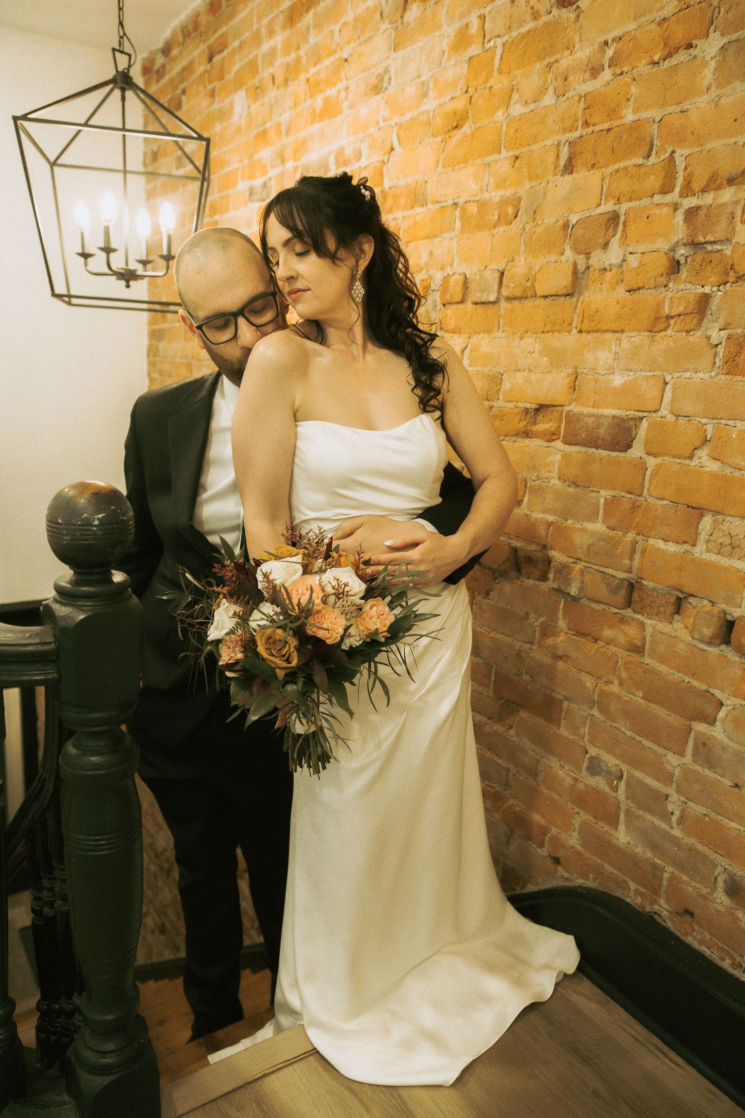 Bride in white satin gown holds floral bouquet as groom in dark suit embraces her near brick wall and chandelier.