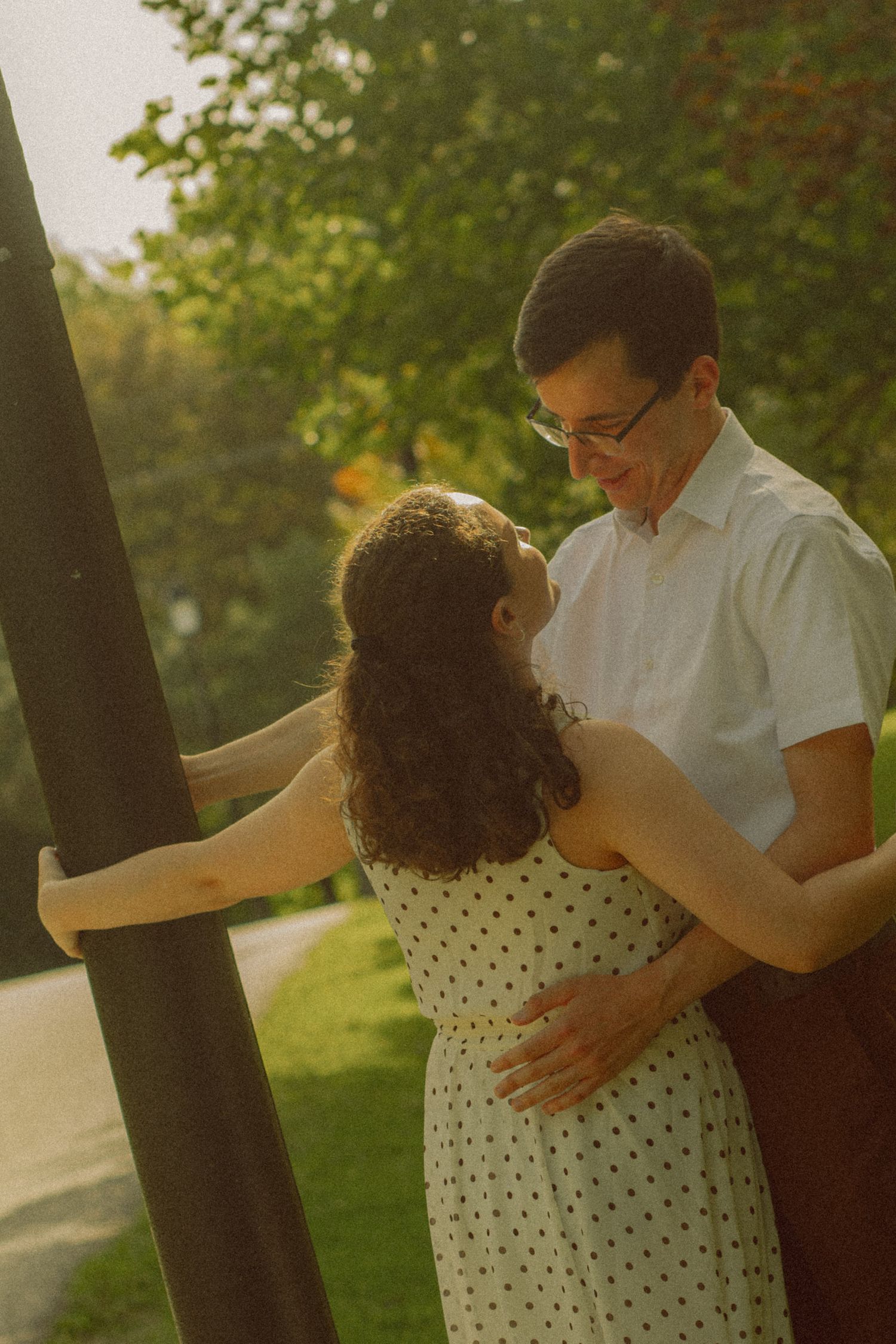 Couple sharing a tender embrace outdoors in warm golden light, woman in polka dot dress, man in white shirt wearing glasses.