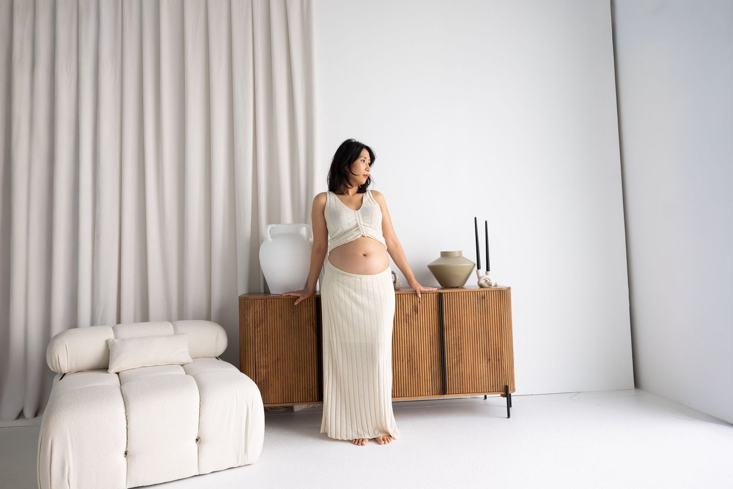Pregnant woman in white crop top and ribbed skirt poses by wooden sideboard in minimalist white studio.