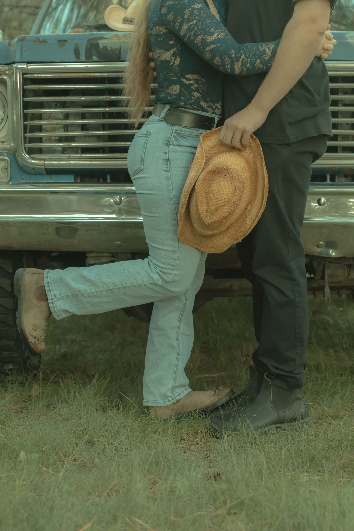 Couple embracing in front of a vintage truck, man holding a cowboy hat, woman in jeans lifting one foot.