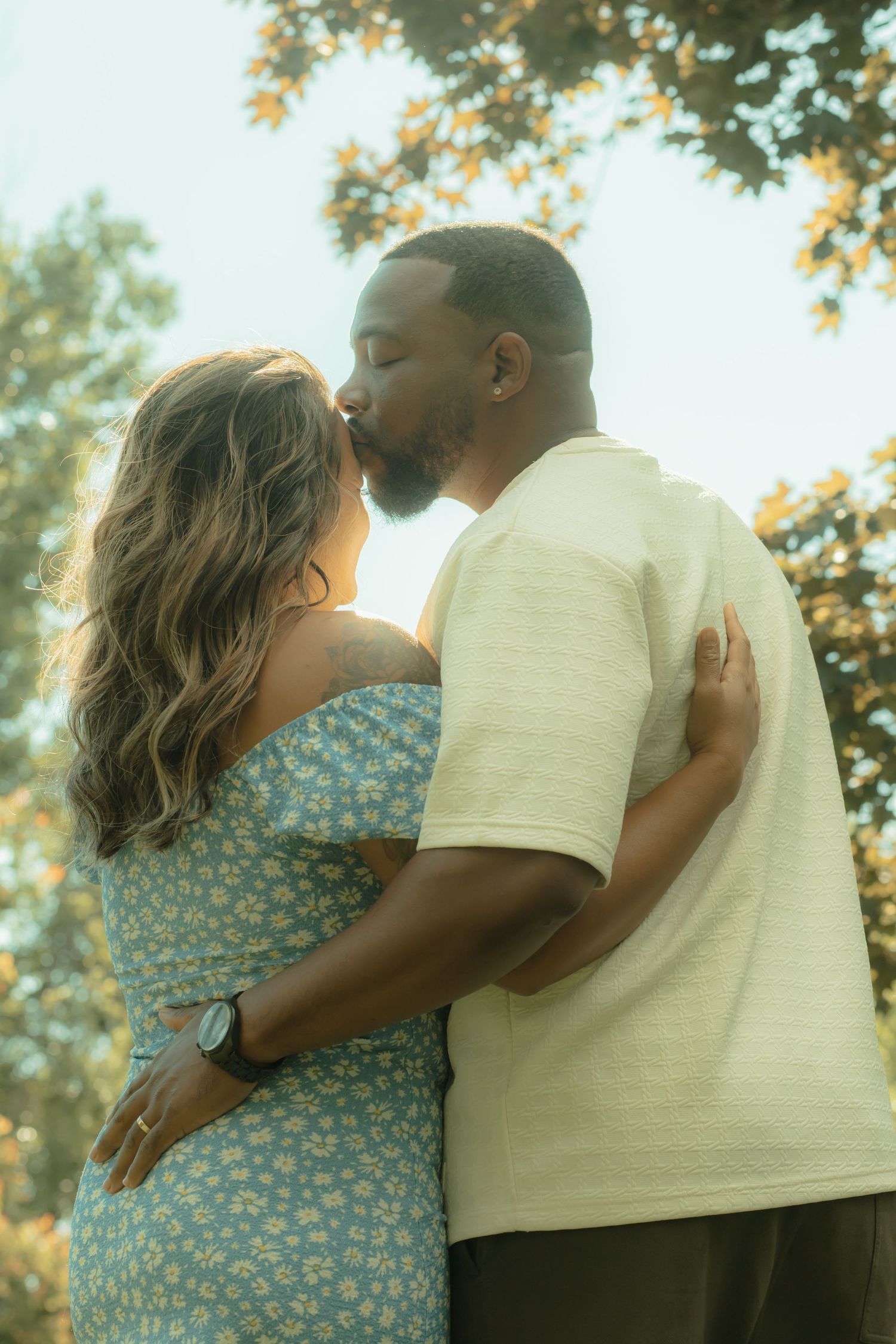 A couple sharing a romantic kiss outdoors, surrounded by lush green trees in warm sunlight.