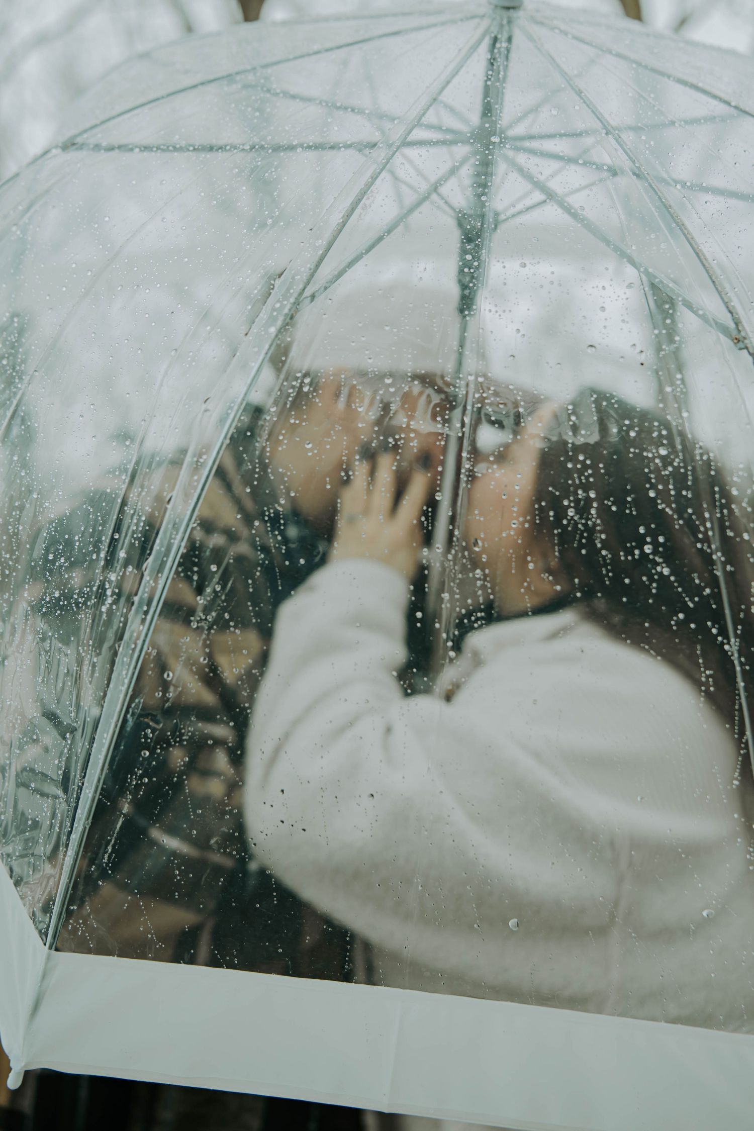 A person in a white sweater looks through a rainy window with water droplets creating a dreamy, atmospheric effect.
