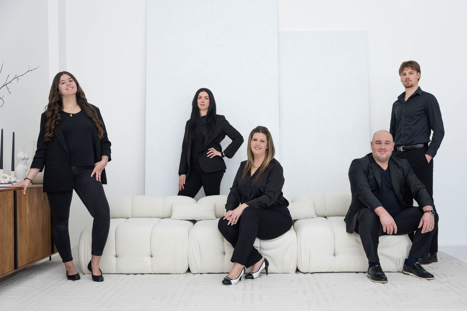 People wearing black business attire sit in various poses against a white wall during a professional photoshoot.