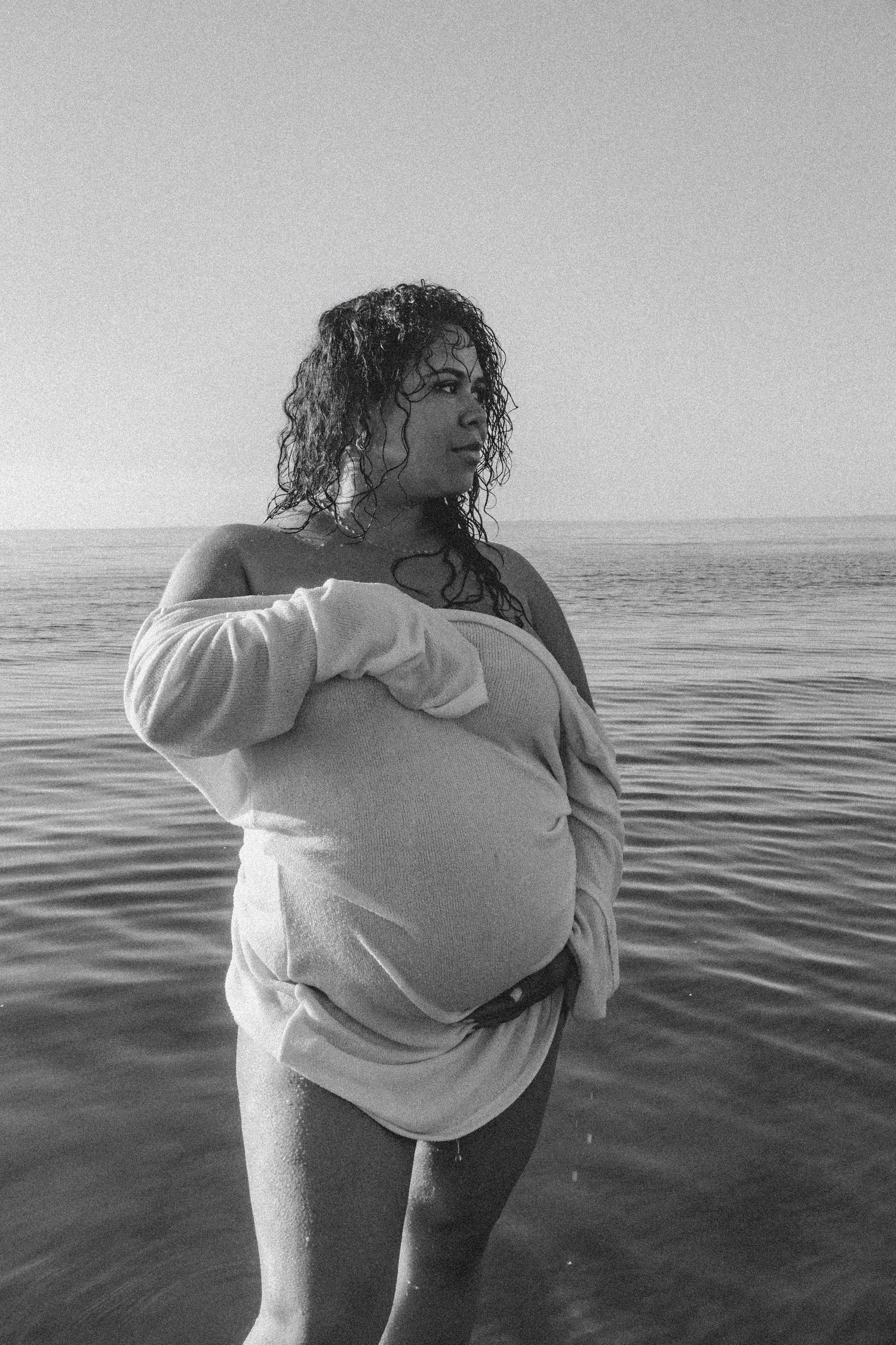 Black and white photo of someone in an oversized sweater standing in ocean waves.