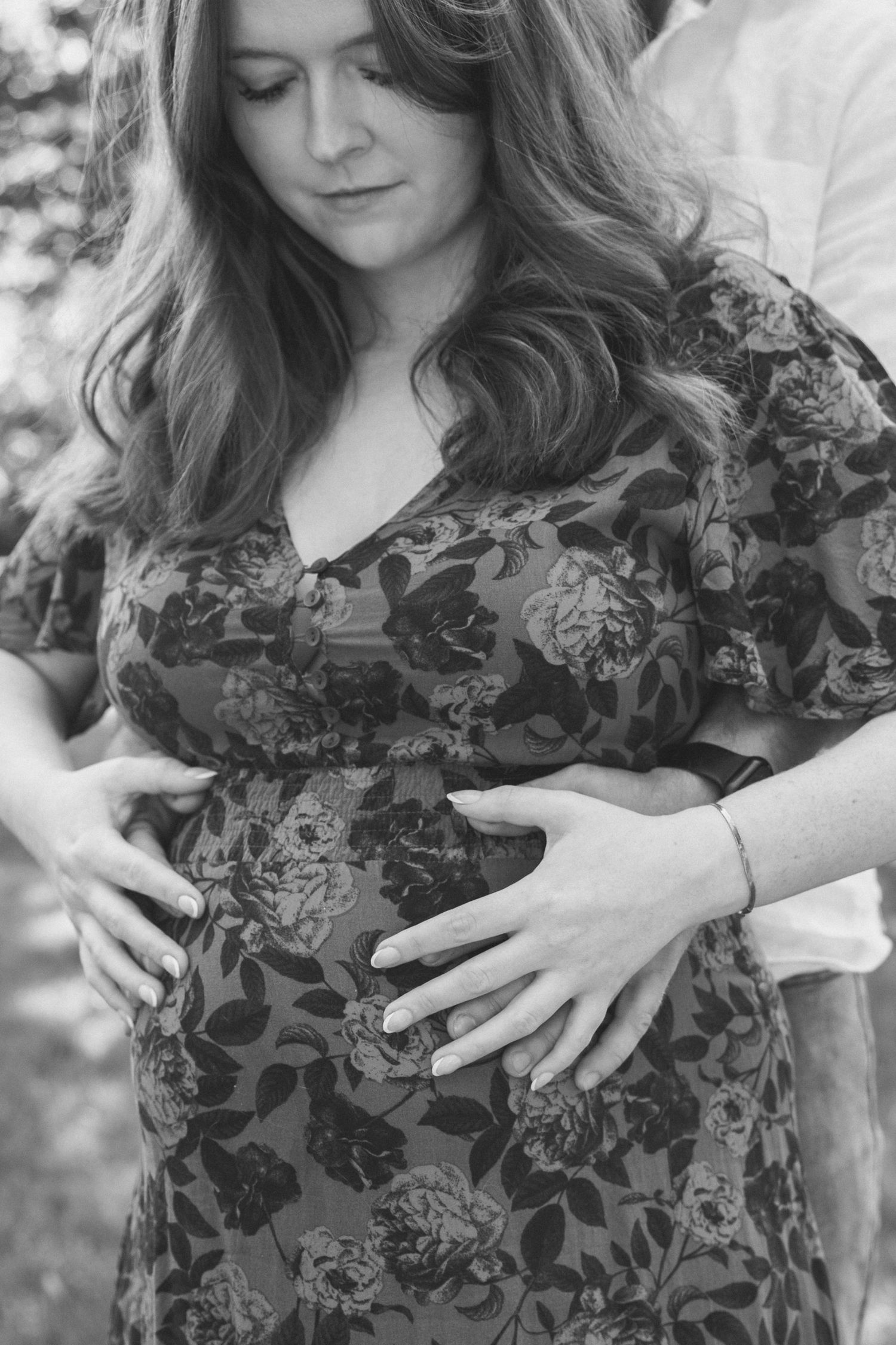 Black and white photo of pregnant woman in floral dress cradling baby bump with partner's hands visible.
