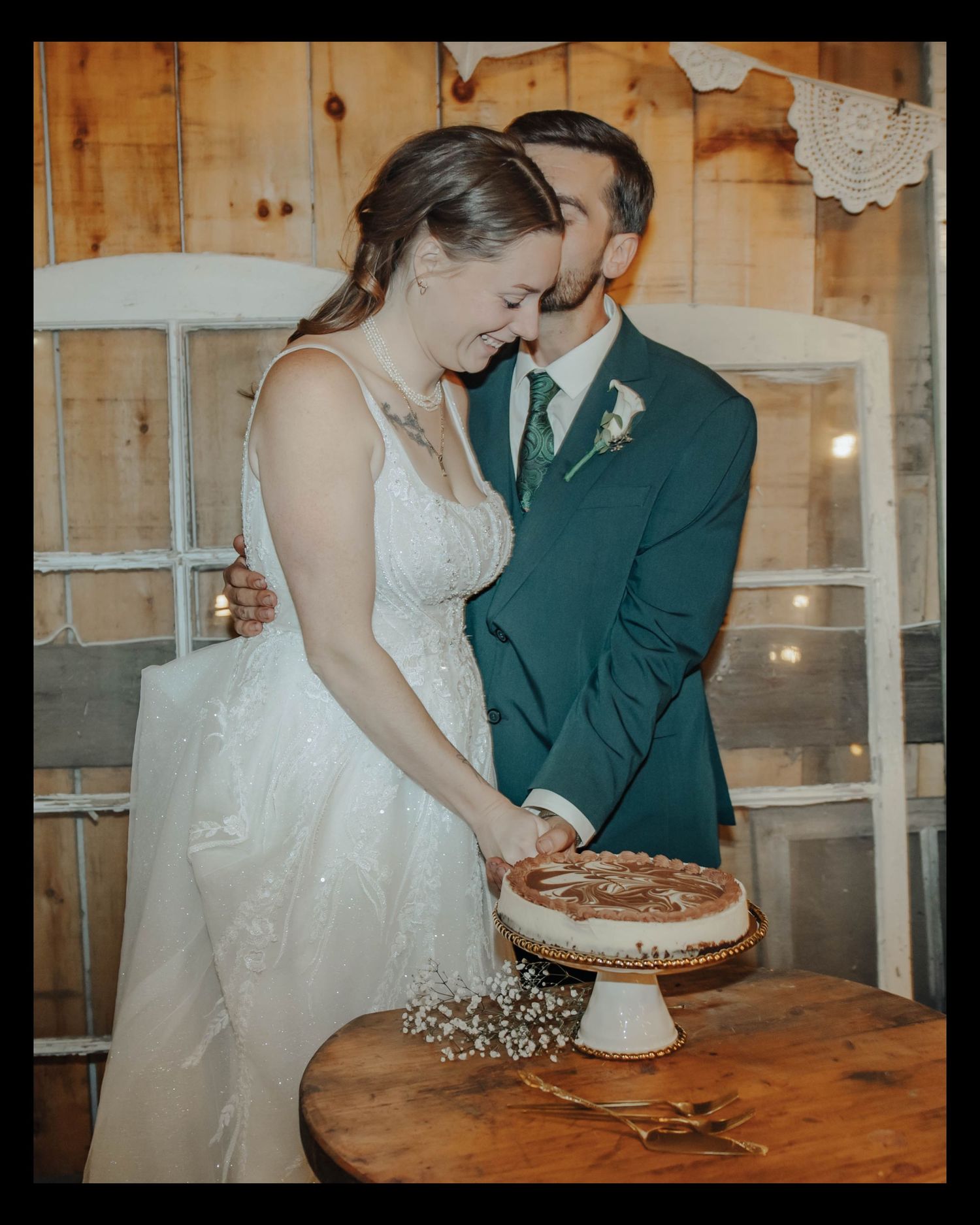 Couple in wedding attire sharing an intimate moment while cutting their rustic naked wedding cake at a barn reception.