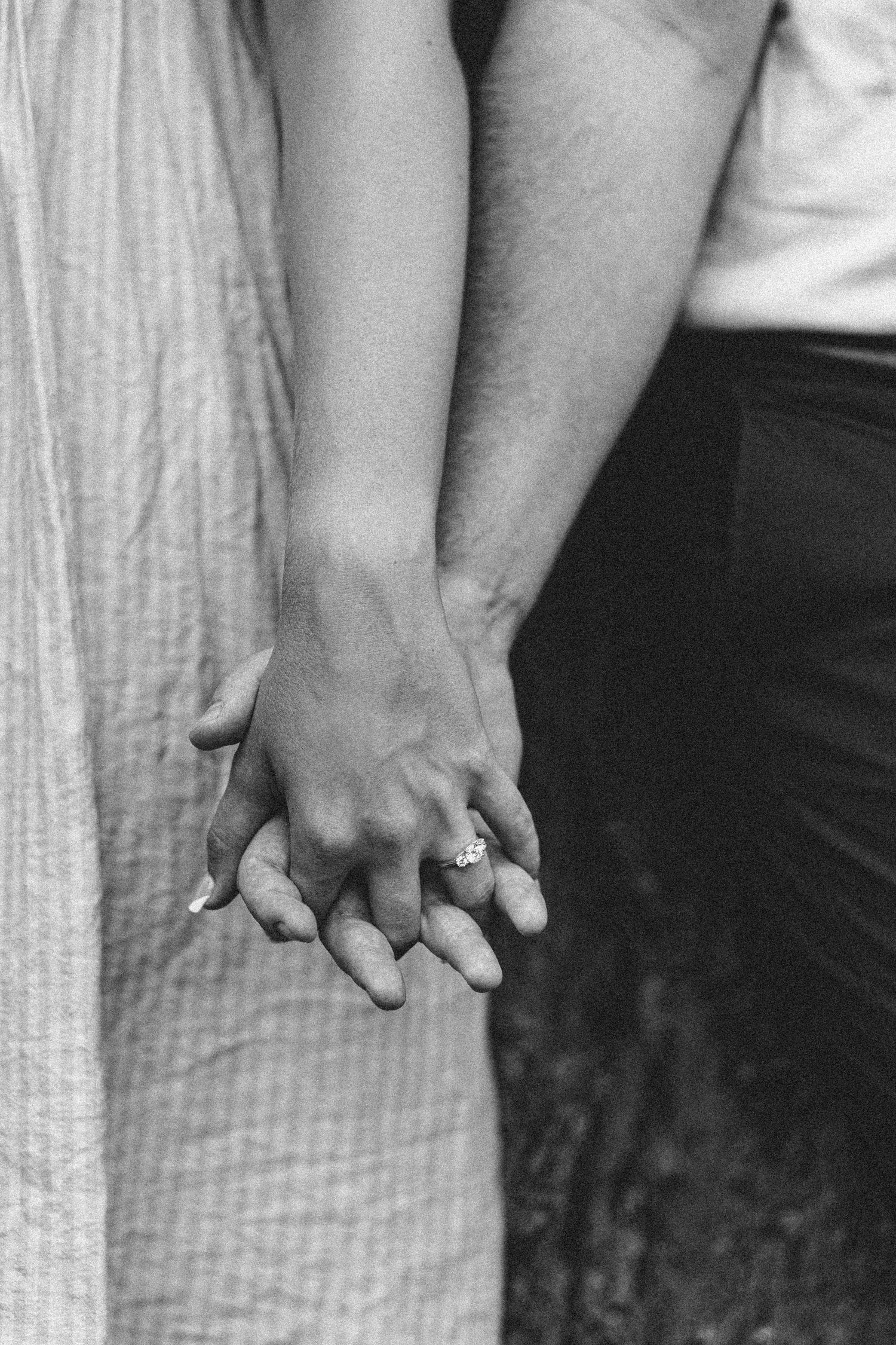 Black and white close-up photograph of two people holding hands with an engagement ring visible.