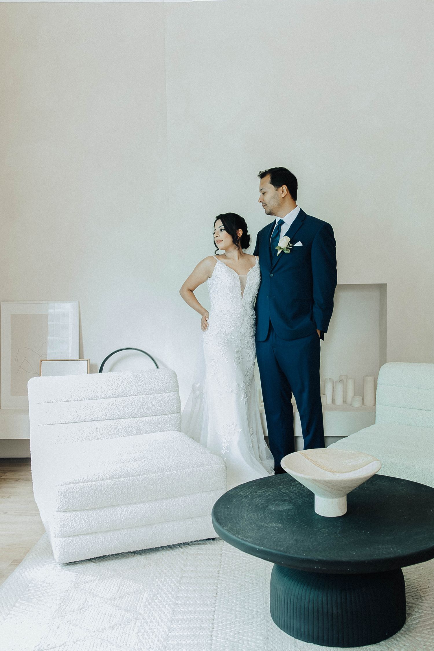 A couple in wedding attire poses together in a modern room with white furniture and a dark round coffee table.