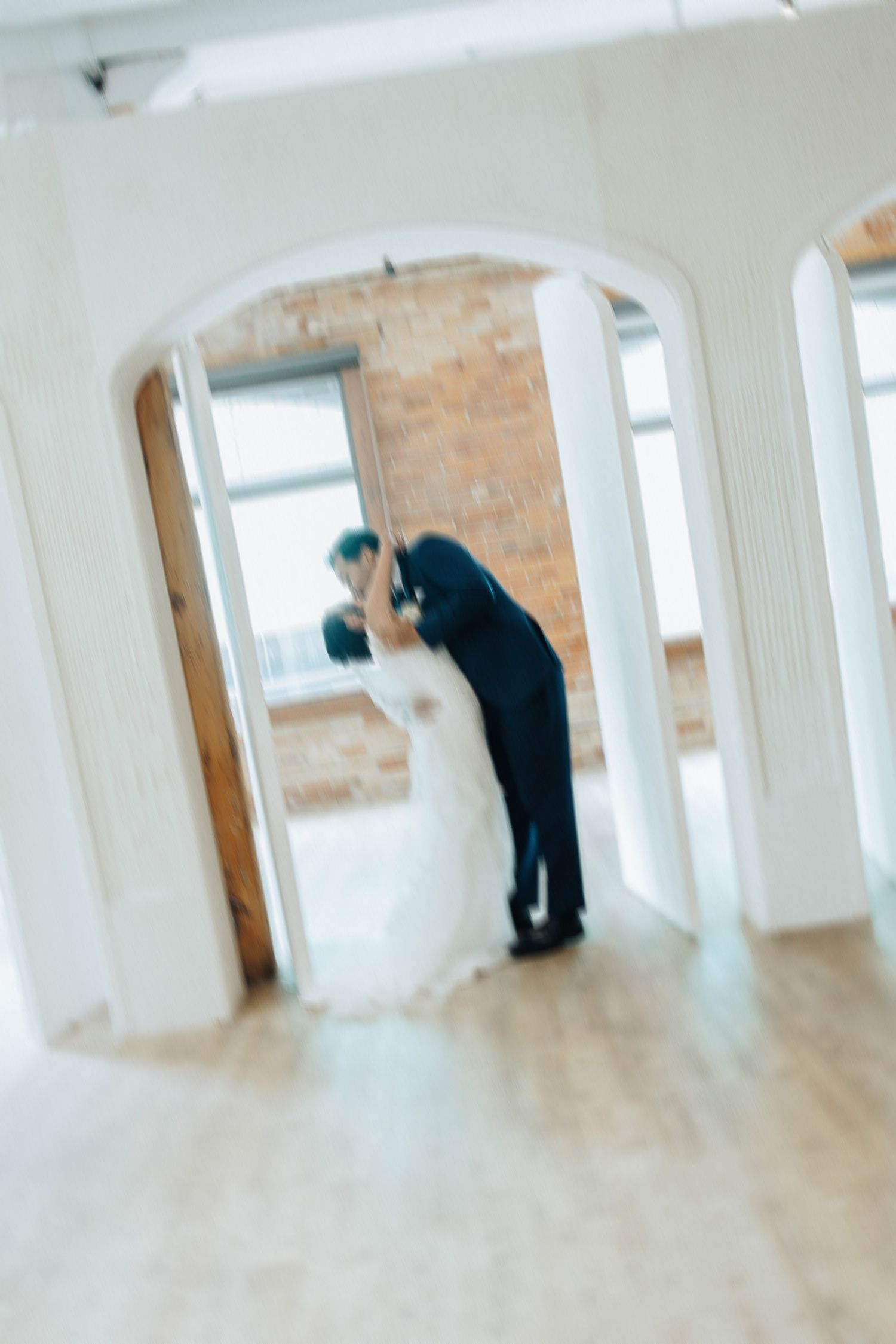 A dreamy wedding moment captured through a doorway showing a couple in white and black sharing a tender kiss on a dance floor.