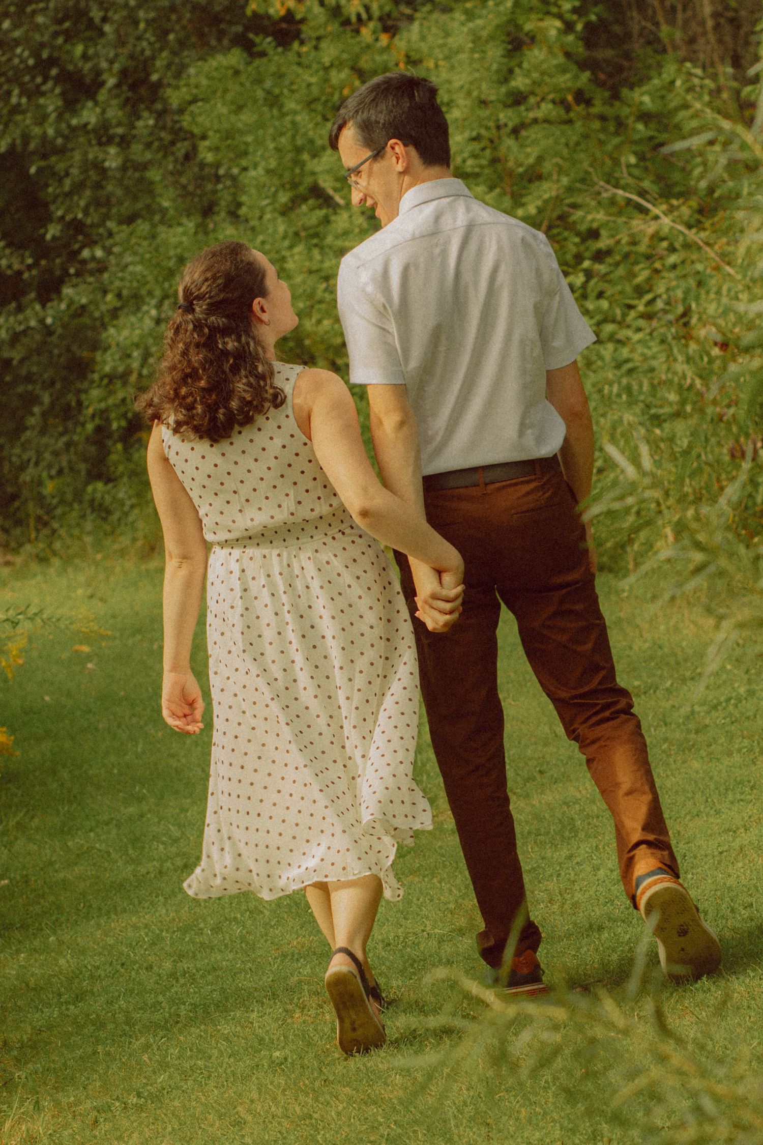 Couple walking hand in hand through lush green garden, woman in polka dot dress, man in white shirt and brown pants.
