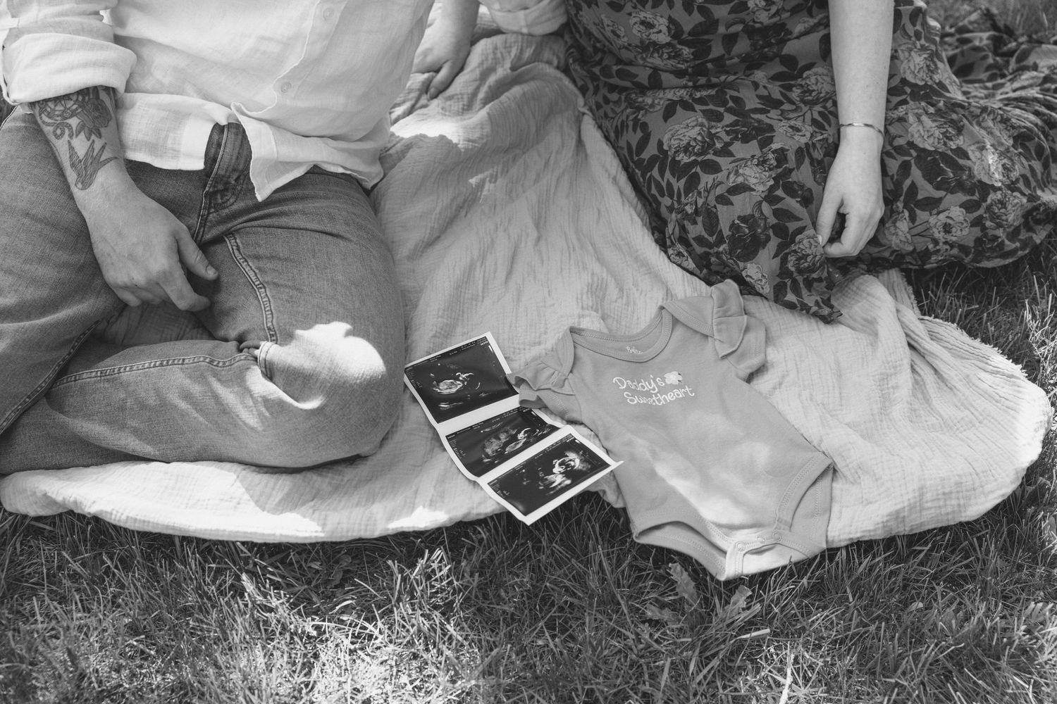 Black and white photo of couple sitting on grass holding ultrasound photos, pregnancy announcement.