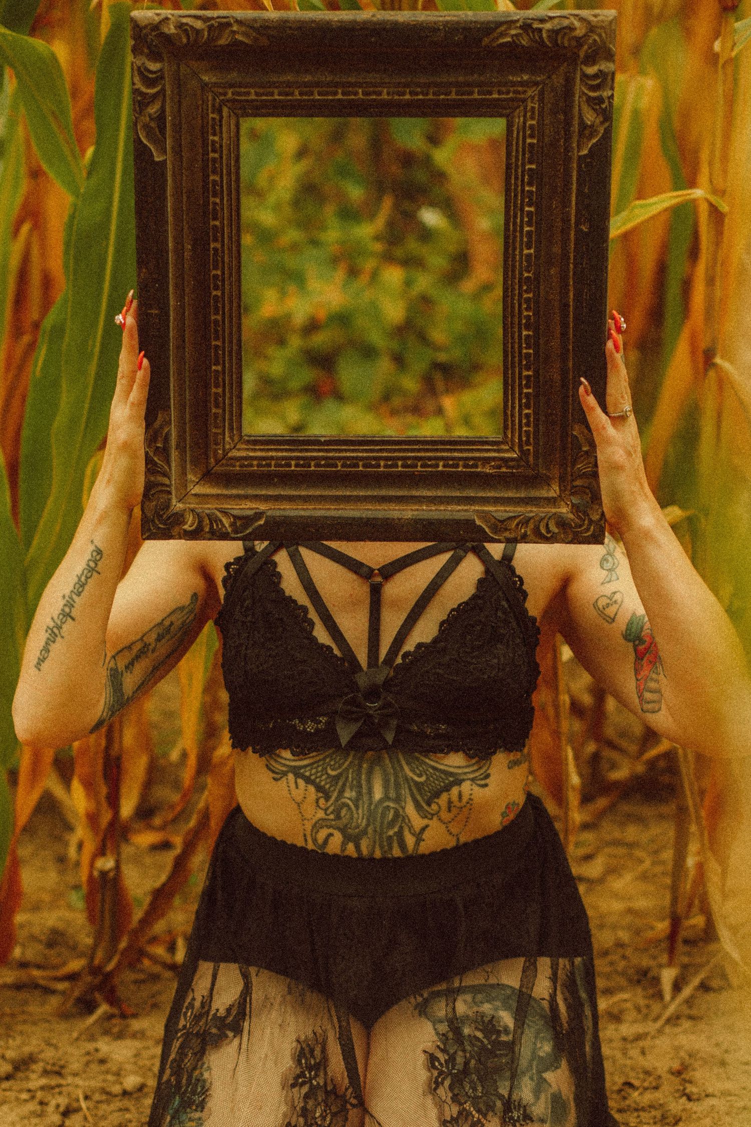 Tattooed woman in black lace bralette holds ornate vintage picture frame over her face outdoors in a corn field.