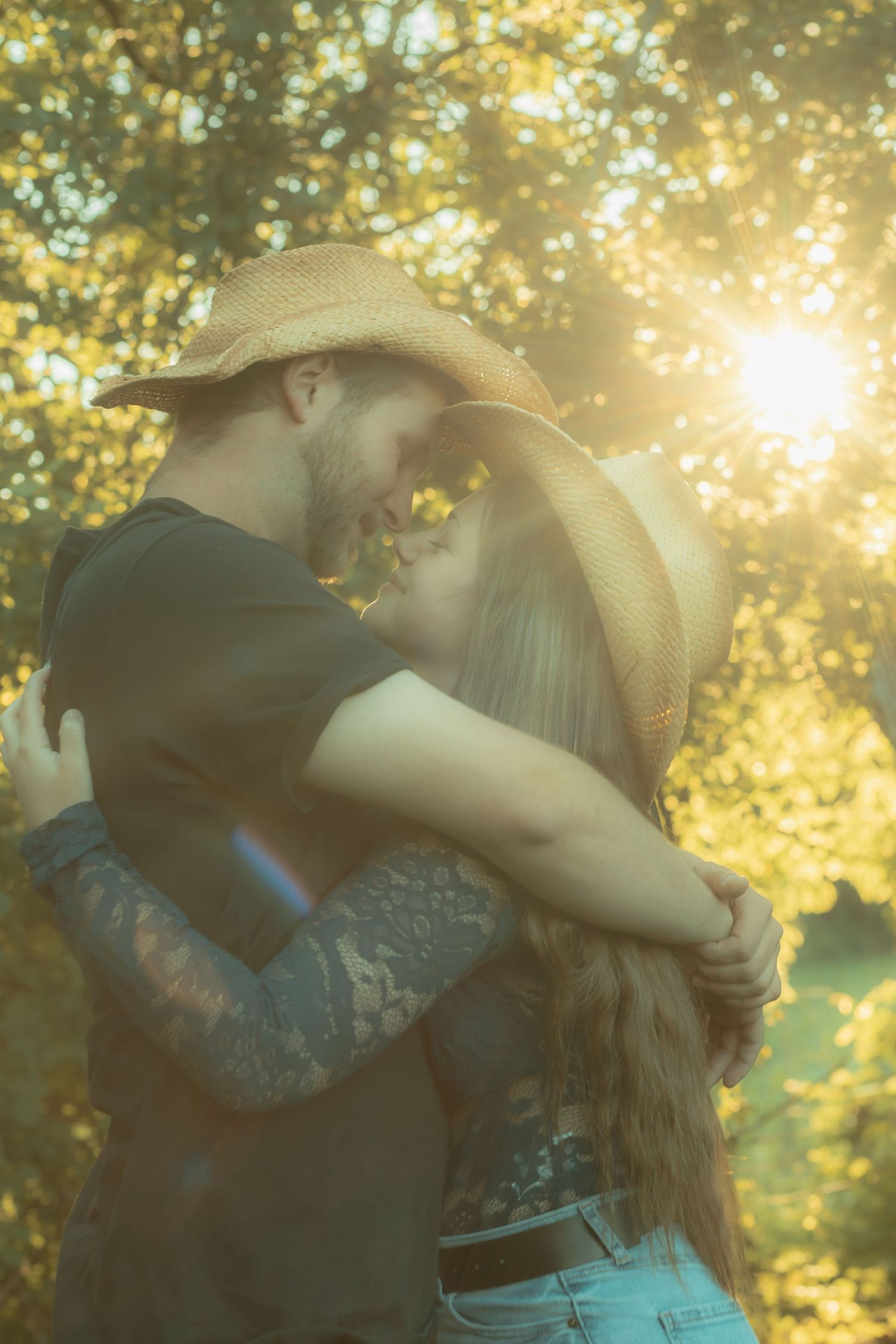 Couple in straw cowboy hats embracing and kissing outdoors in golden sunlight with trees in background.