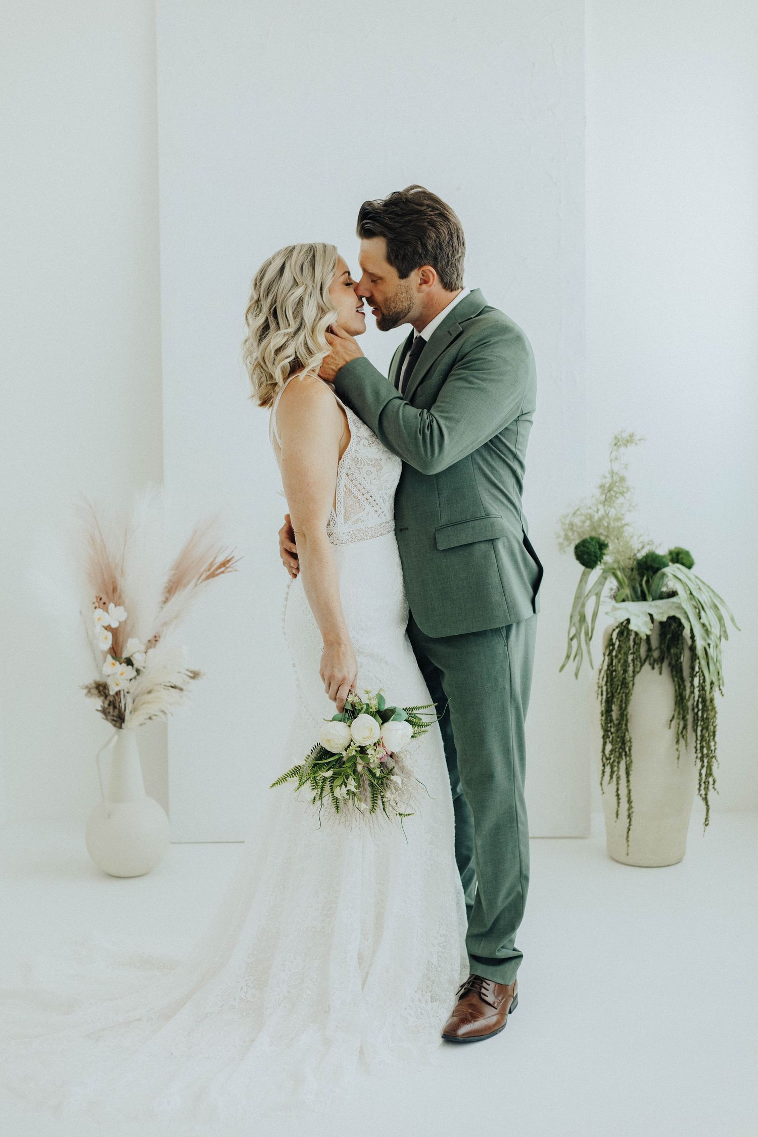 A romantic wedding couple shares a tender moment in a minimalist white room decorated with pampas grass and flowers.