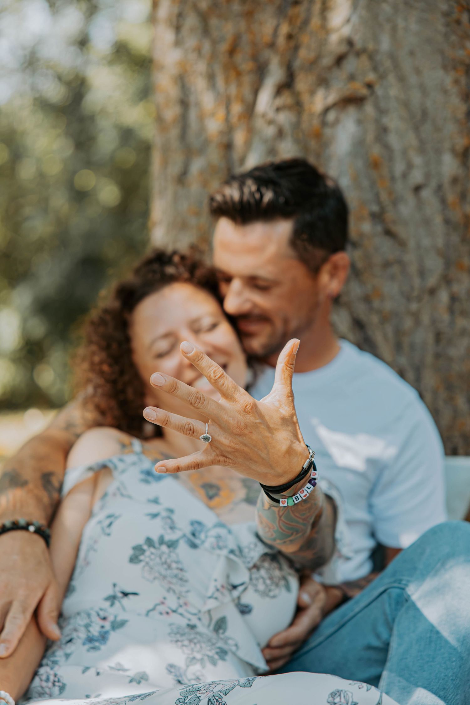 A candid outdoor engagement photo showing a couple laughing together while displaying a ring in the sunlight.
