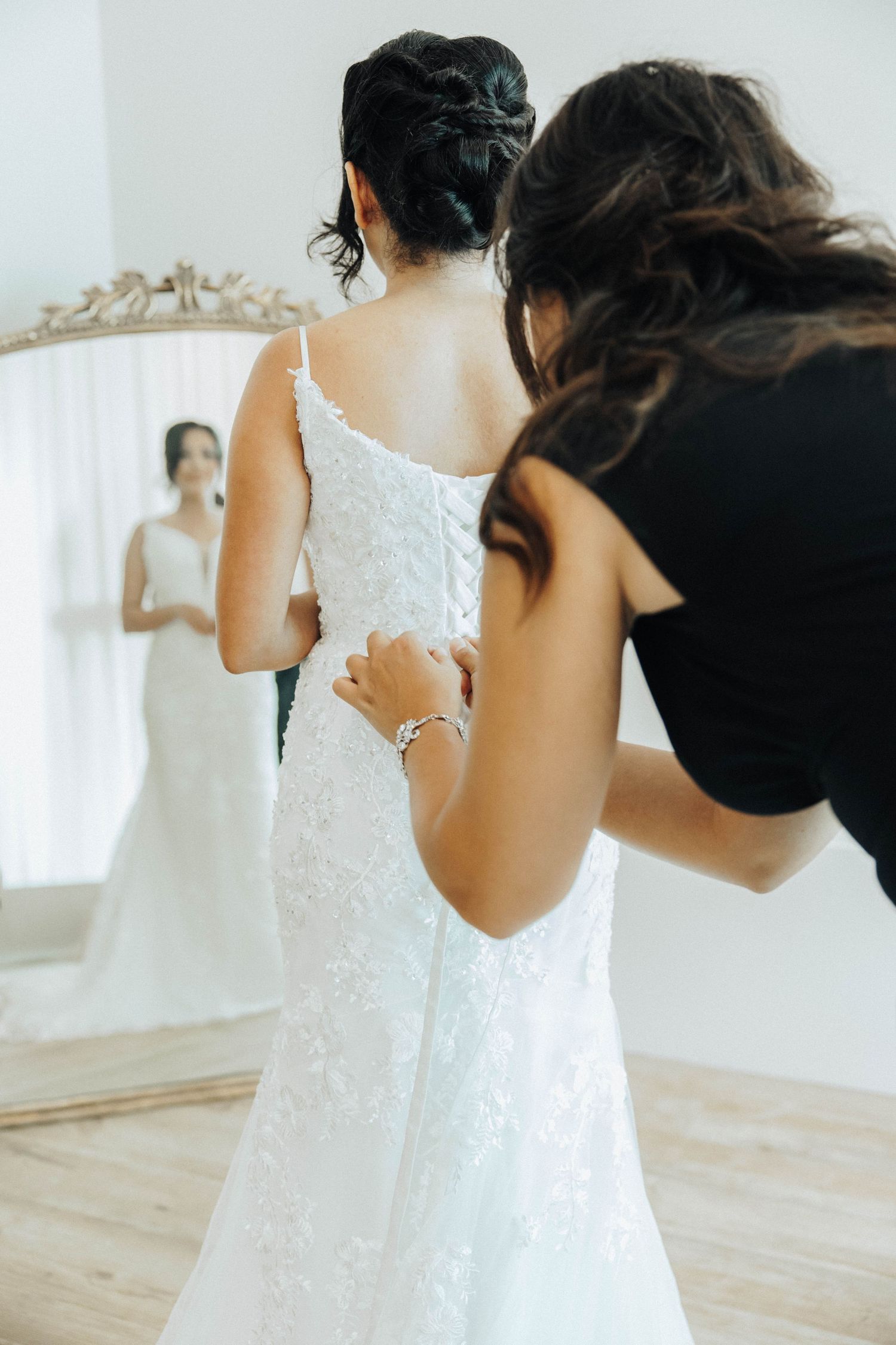 Someone helps zip up a white lace wedding dress while standing in a bright room with hardwood floors and a mirror.