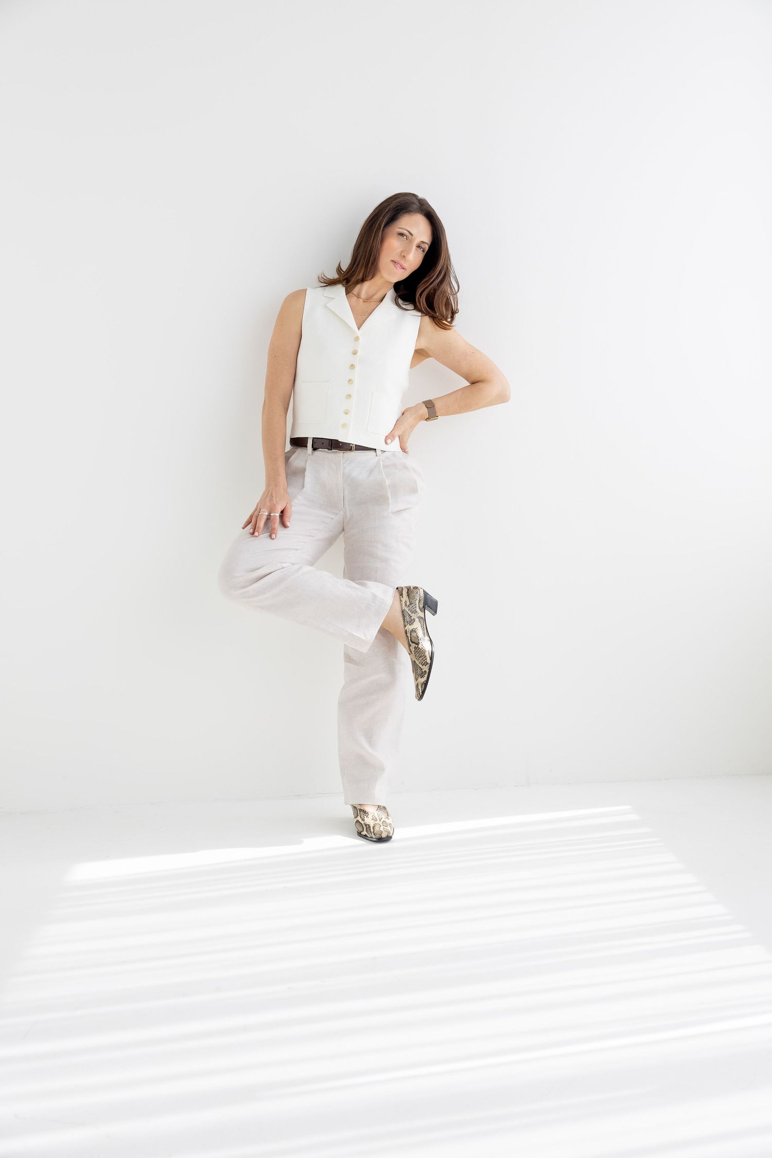 Woman in white sleeveless vest and pants posing against white wall wearing snakeskin heels.