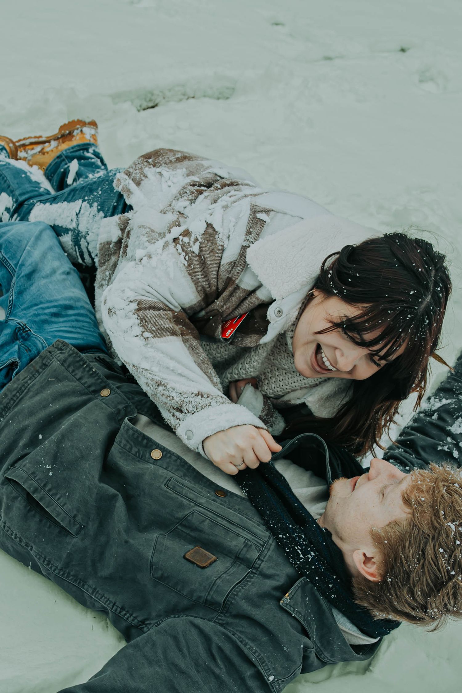 Two people lying in snow wearing winter coats share a tender moment together.
