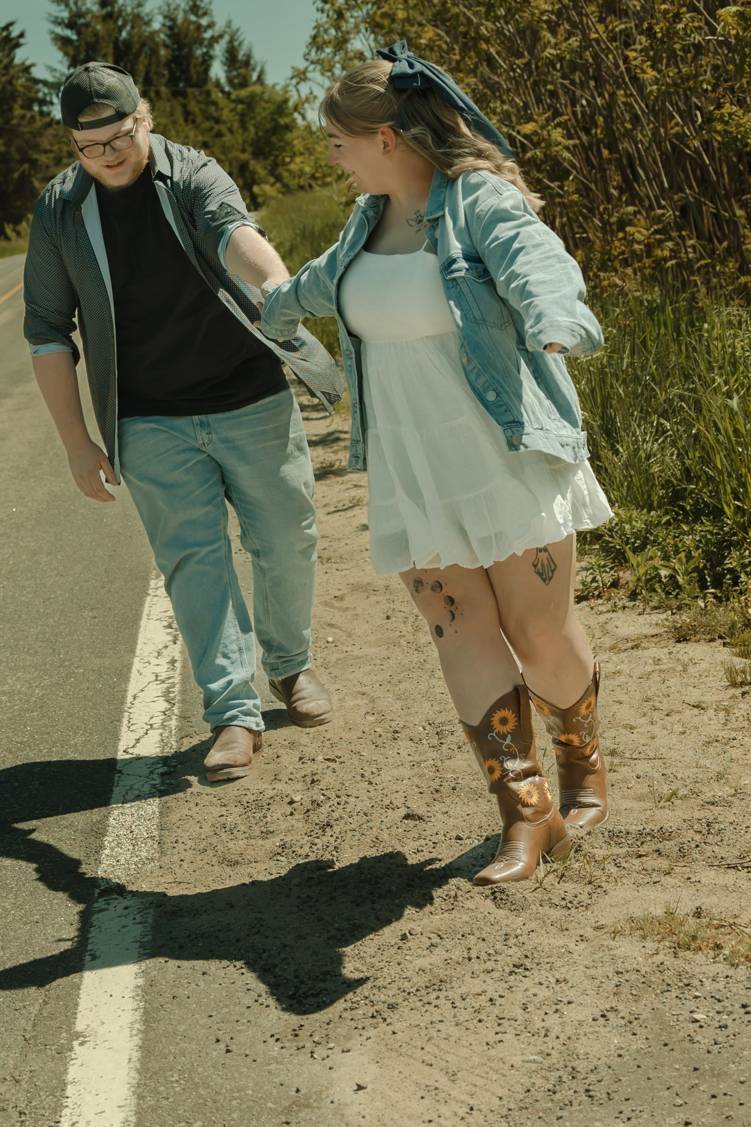 Couple walking along a sunny roadside, woman in white dress, denim jacket, and cowboy boots holding hands with man in jeans.