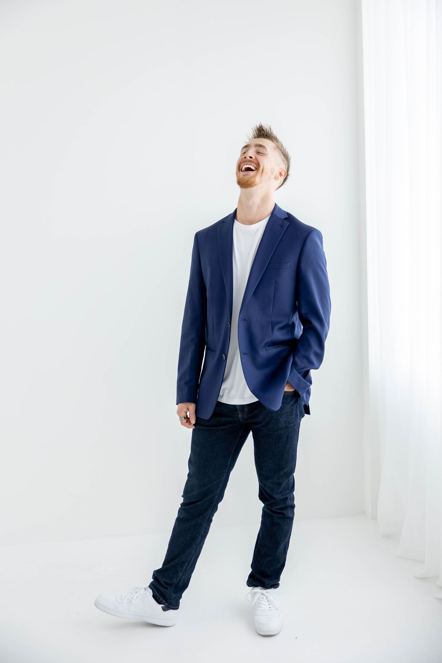 Person wearing navy blazer and white sneakers stands casually against white studio background.