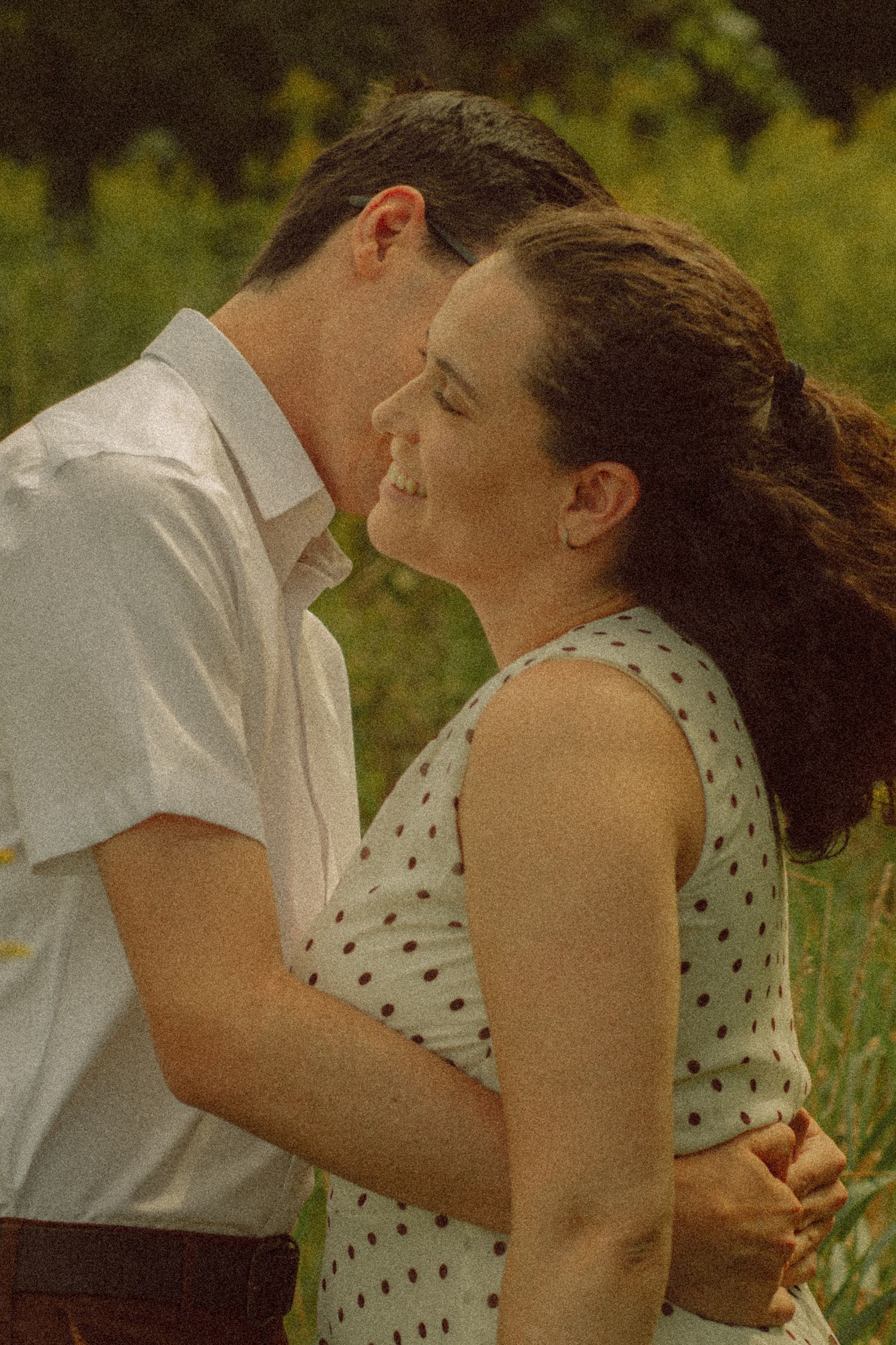 Romantic couple sharing a tender moment outdoors, woman in polka dot dress smiling as man leans in for a kiss.
