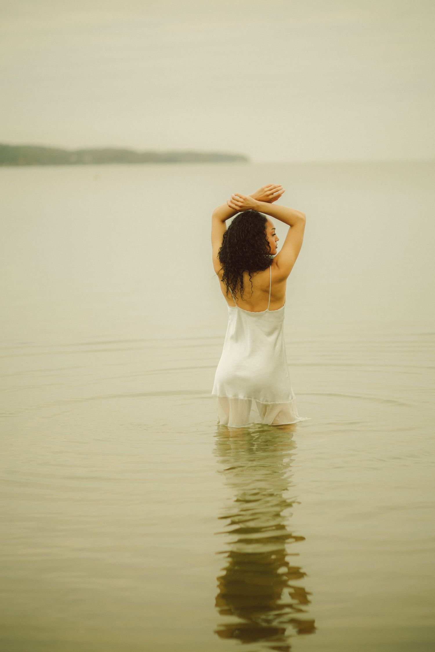 Woman in white slip dress standing waist-deep in calm water, arms raised gracefully overhead, viewed from behind.