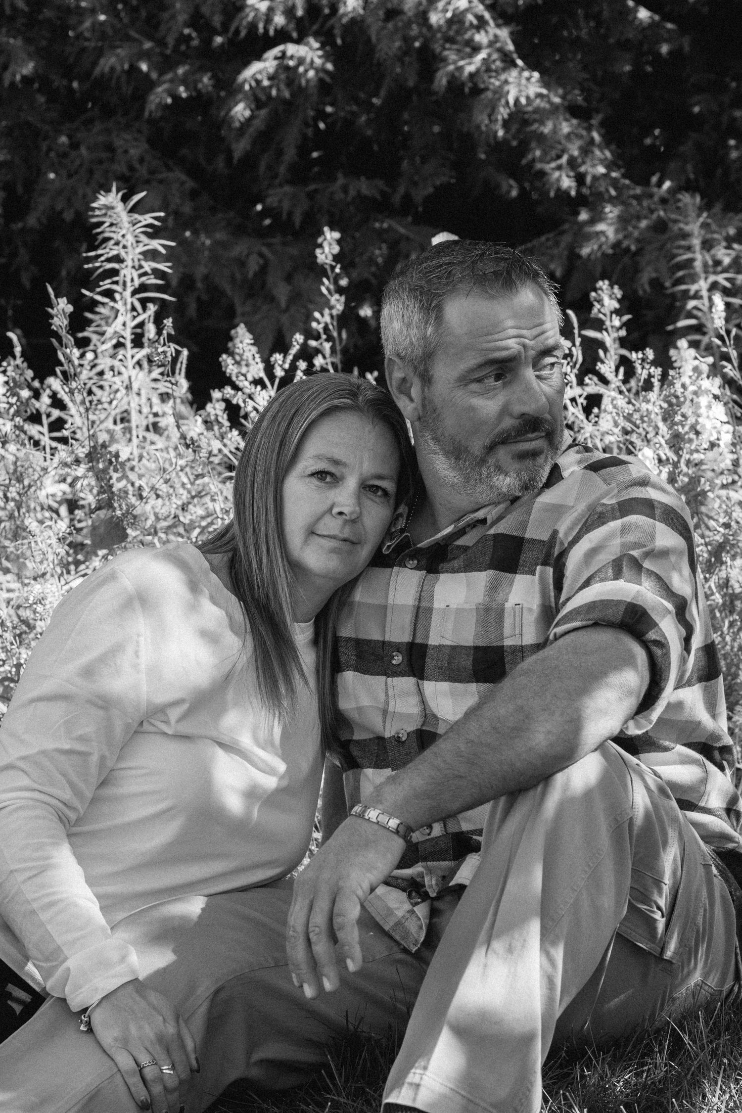 Black and white photo of a middle-aged couple sitting together outdoors, leaning close with trees and plants in background.