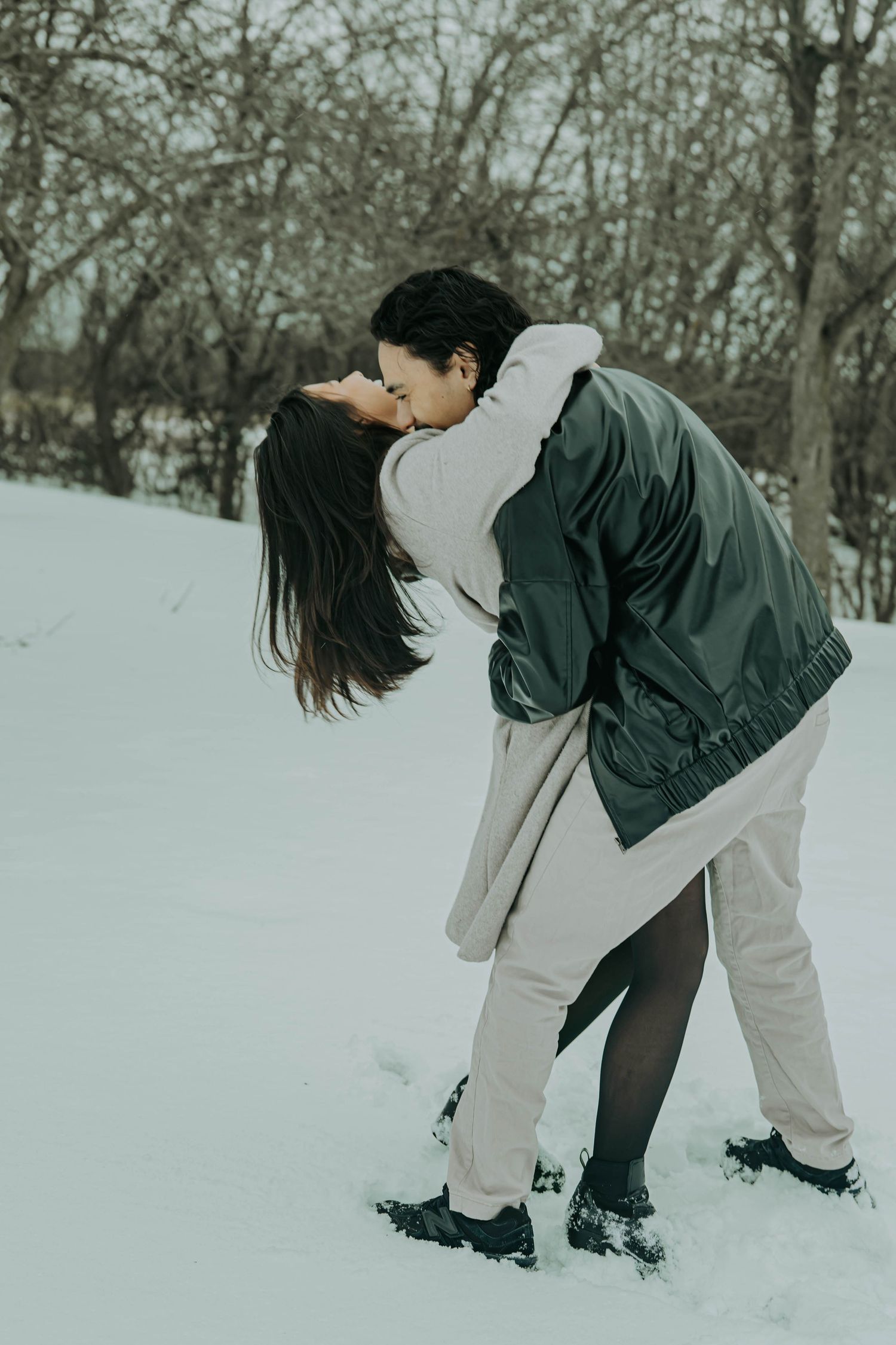 A romantic winter scene shows a couple in warm clothing embracing in the snow surrounded by bare trees.