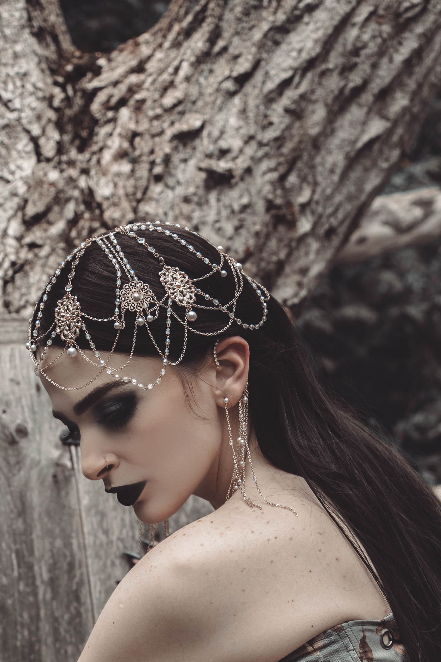 A dark and ethereal photoshoot featuring delicate silver headpieces with intricate beadwork against a textured tree backdrop.