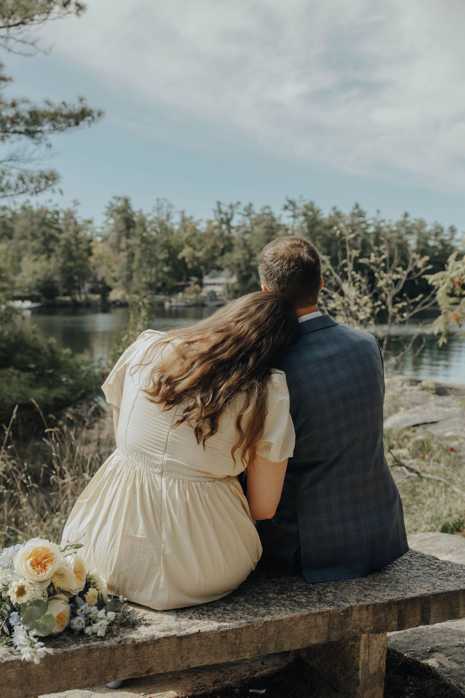 A couple sits on a wooden bench by a tranquil lake surrounded by trees, creating a romantic lakeside moment.