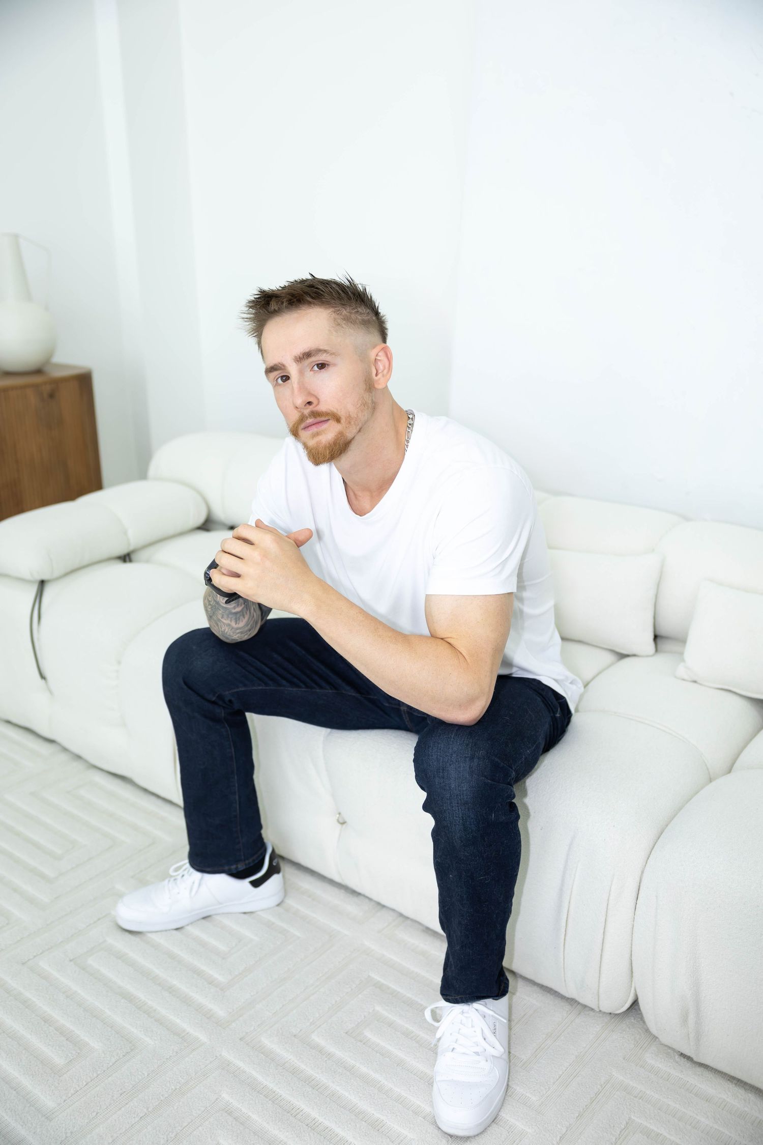 A person in a white t-shirt and dark pants sits contemplatively on a white couch in a minimalist room setting.