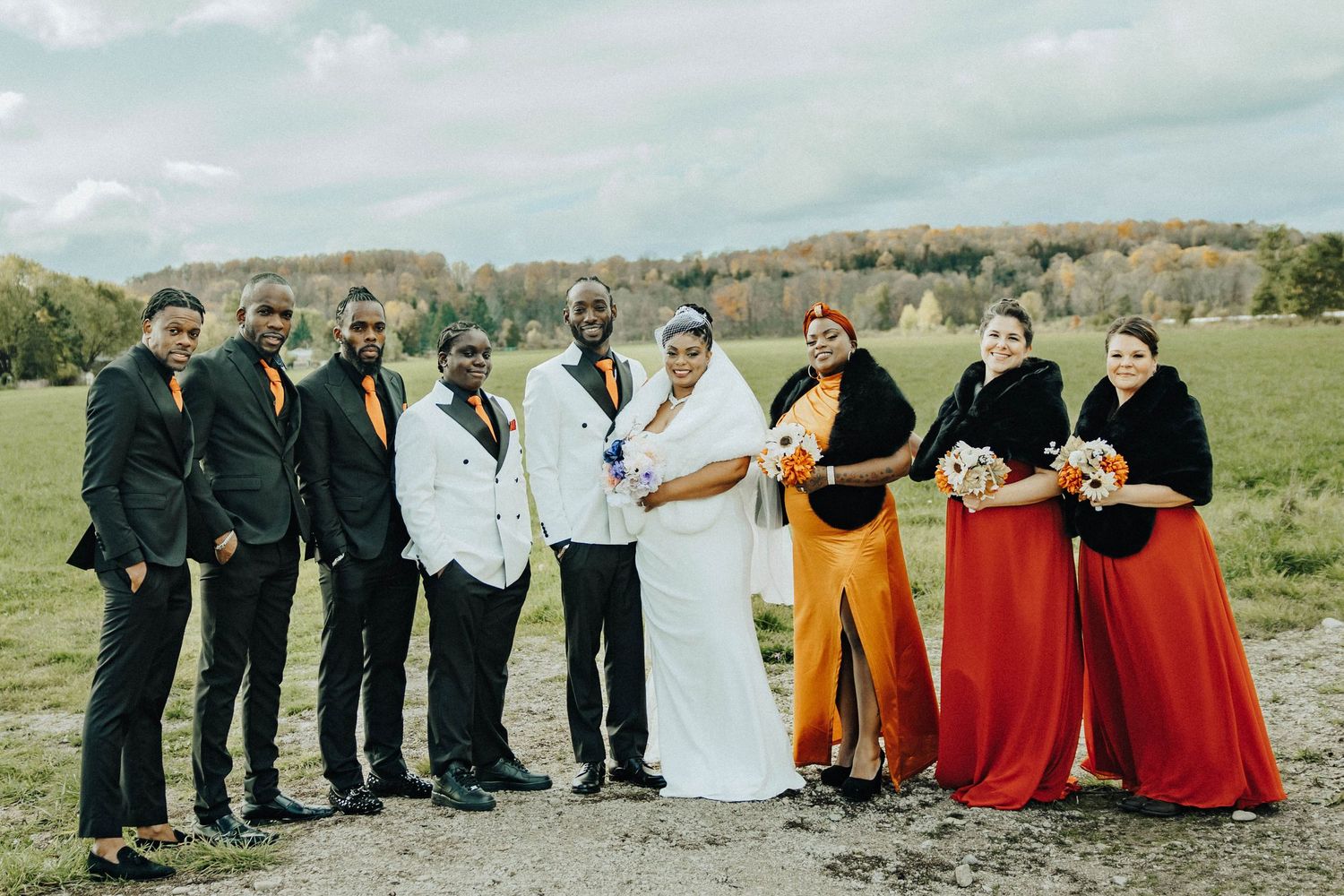 Wedding party poses outdoors in formal attire with bridesmaids in red and orange skirts against a rural backdrop.