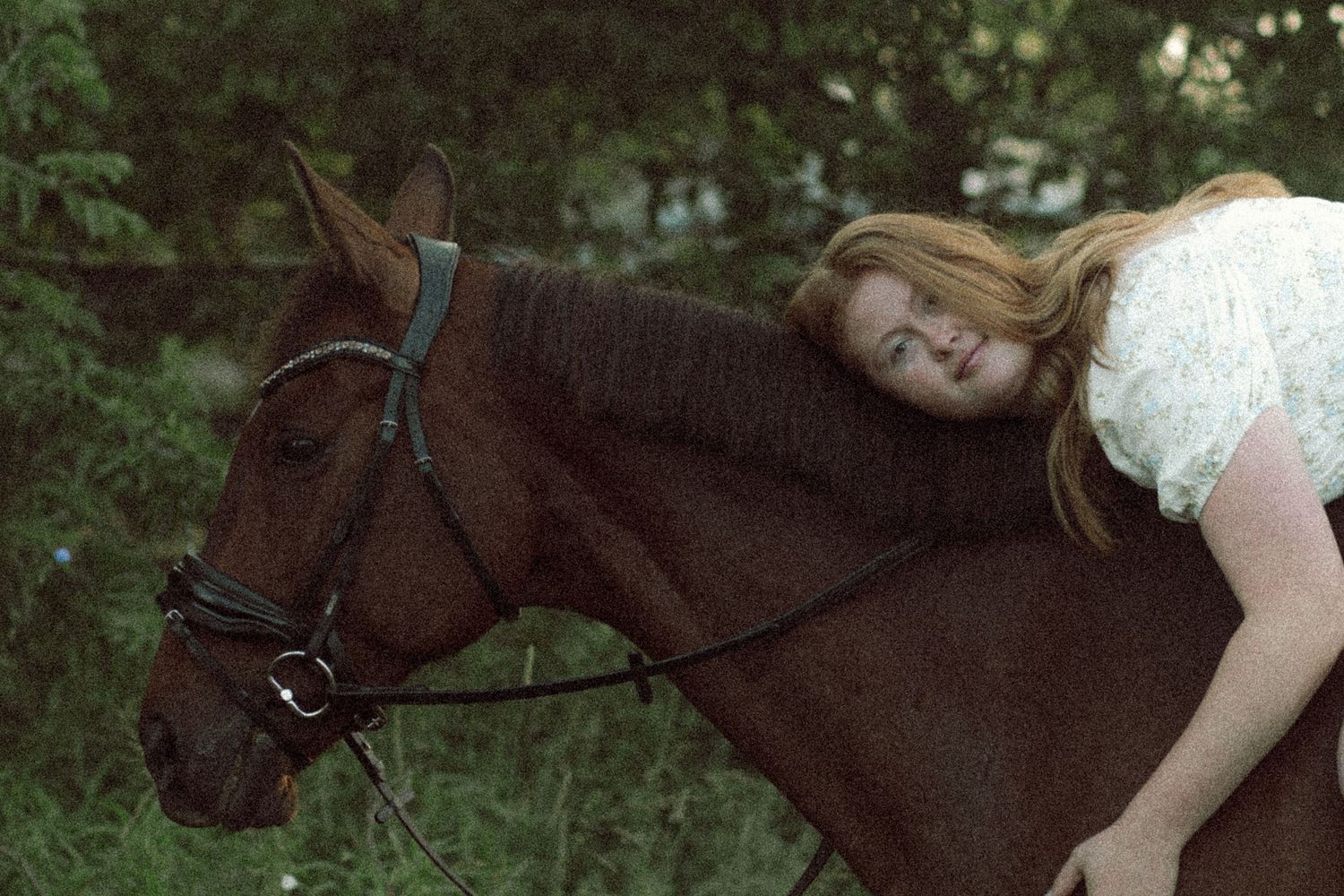 Young woman with red hair leaning forward on a brown horse outdoors near green trees.