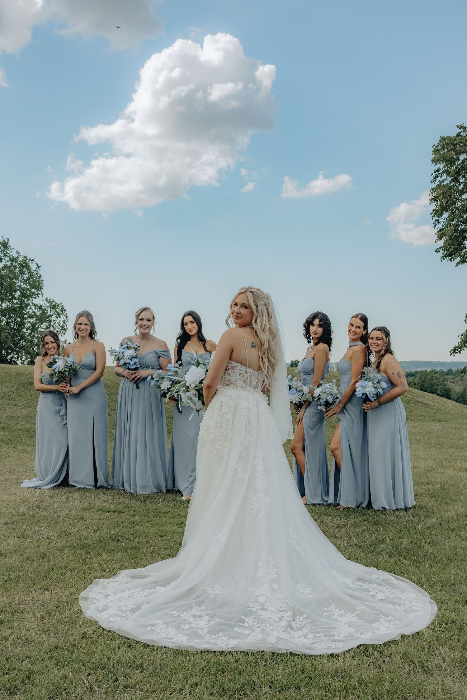 Bride in lace gown with train stands before bridesmaids in dusty blue dresses holding bouquets on green hillside.