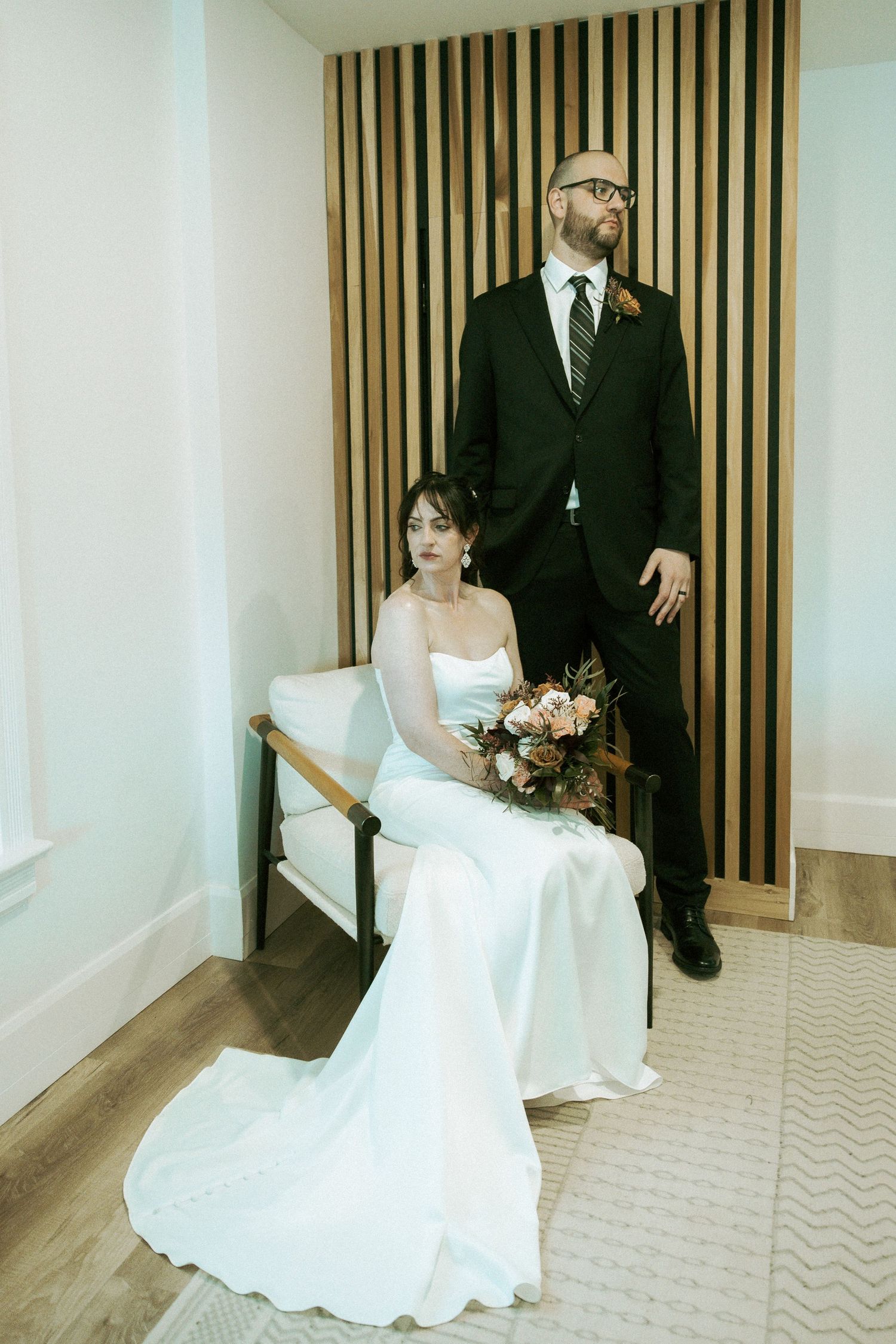Bride sits in chair holding bouquet while groom stands beside her against modern wood panel backdrop.