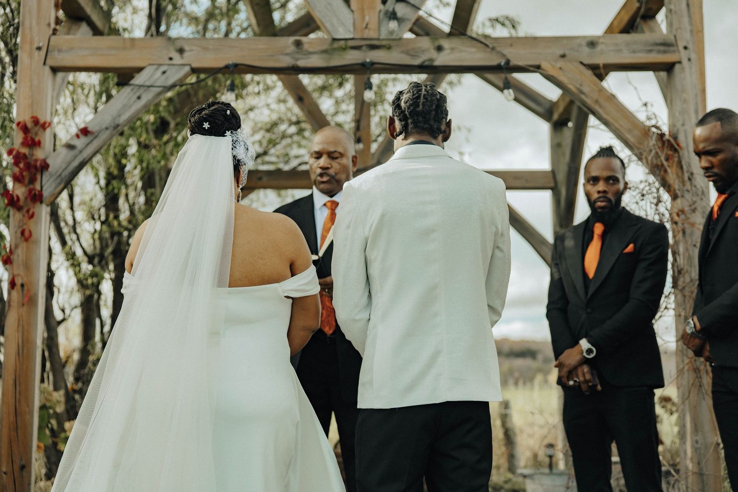 A wedding ceremony takes place under a wooden arch with the couple and officiant standing together during the vows.