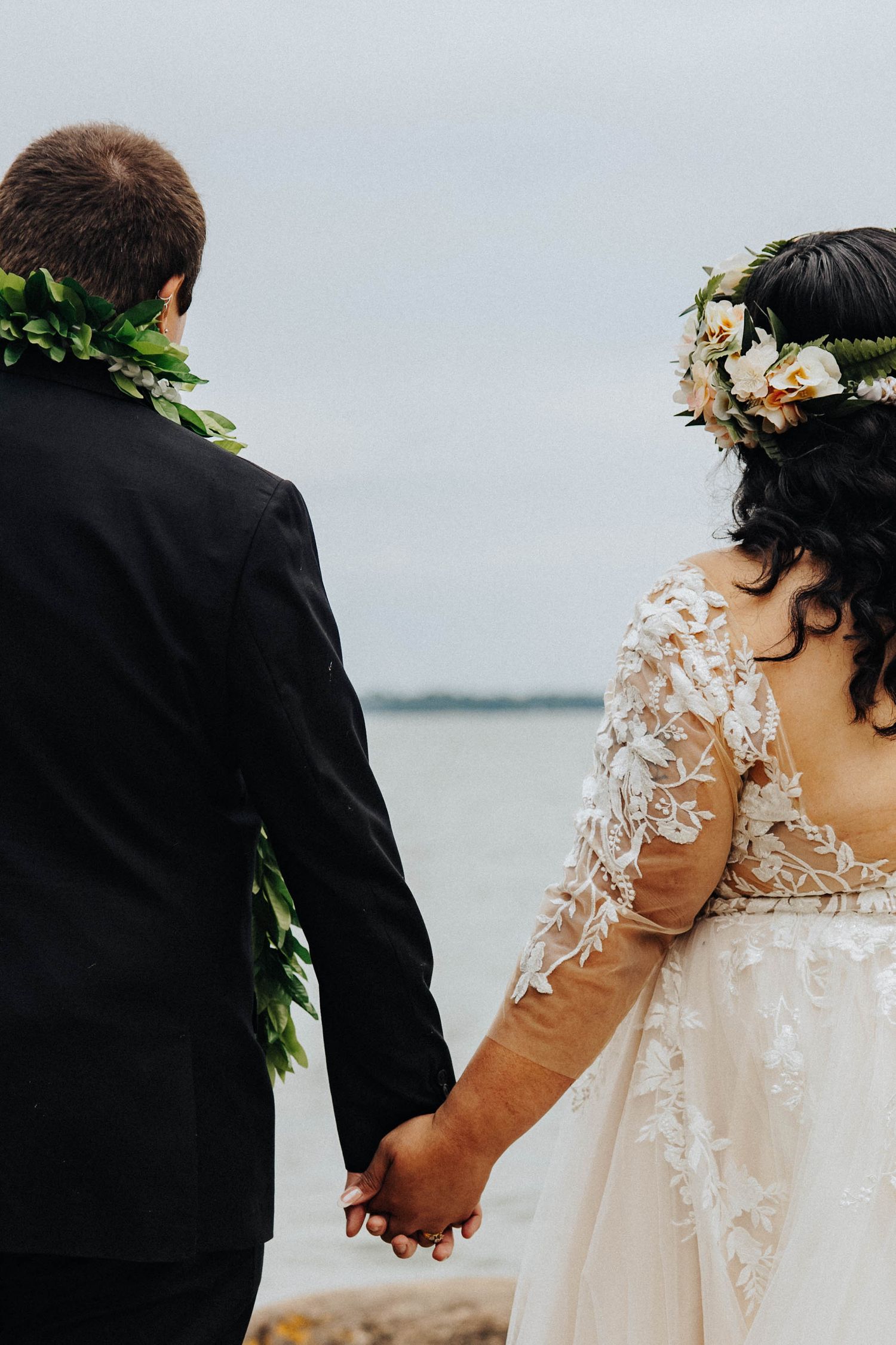 Wedding couple wearing traditional attire with lei and lace dress hold hands by the ocean on their special day.