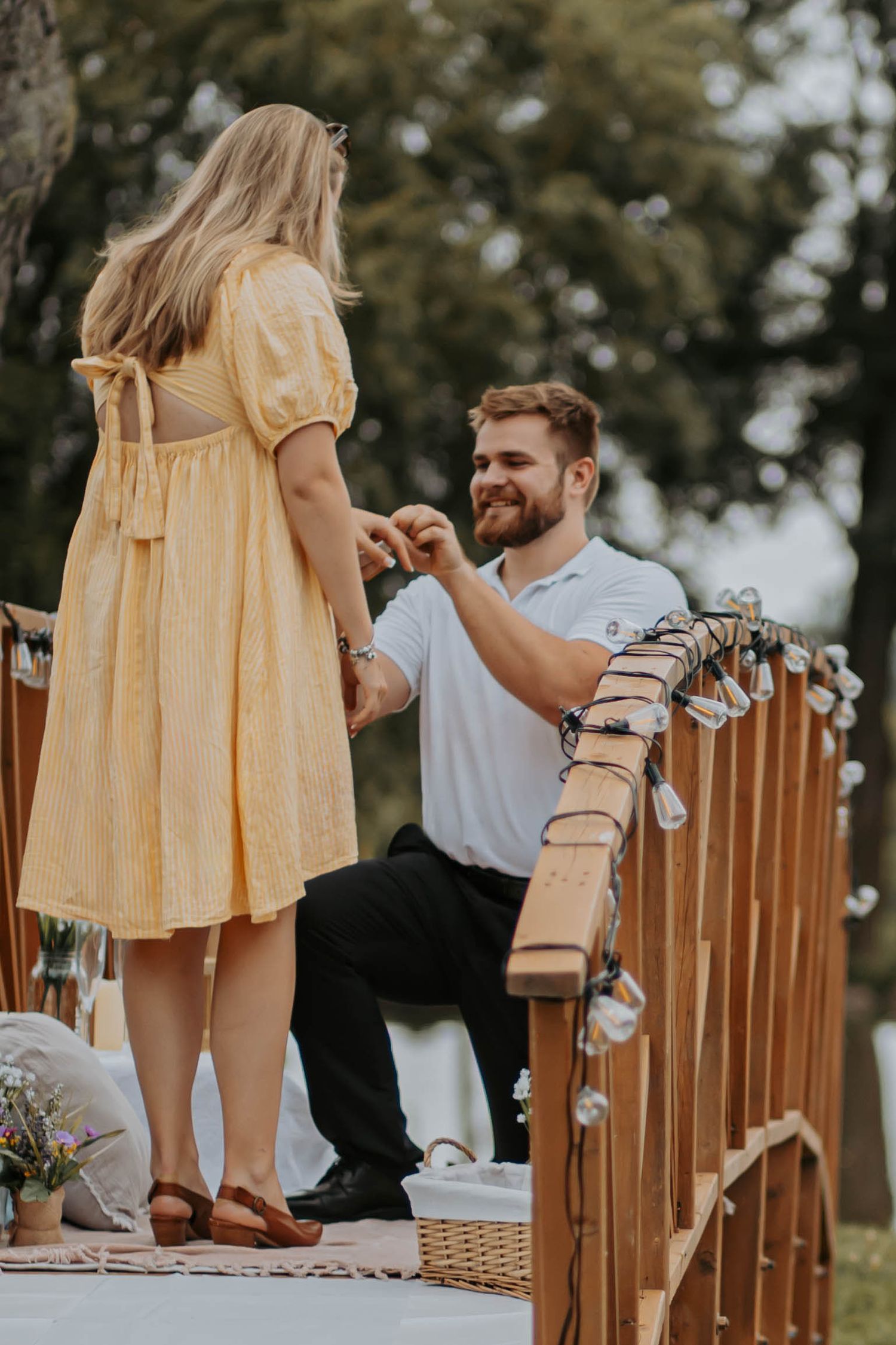 Outdoor engagement scene on a rustic wooden bridge with yellow dress and fairy lights creating romantic atmosphere.