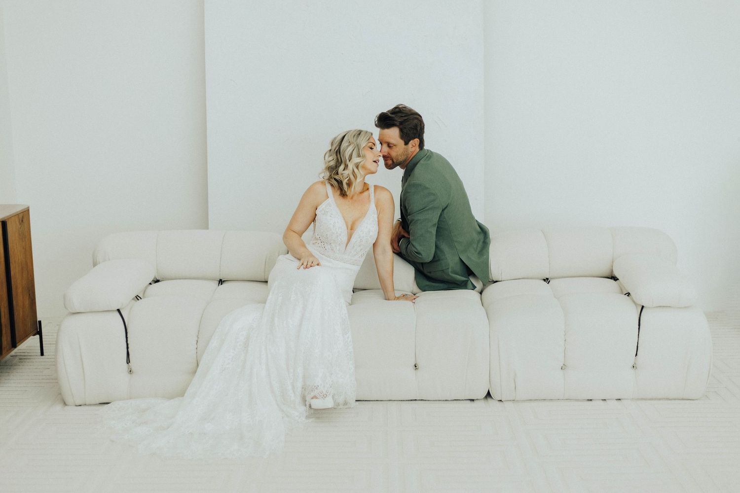 A couple shares a tender moment on a white couch in an elegant minimal setting during their wedding day.
