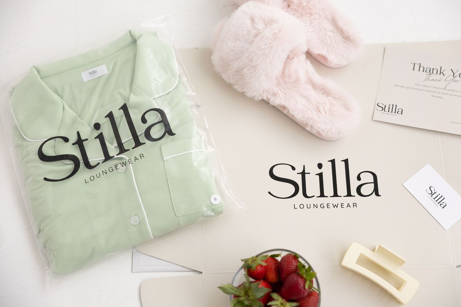 Stilla Loungewear brand flatlay with mint green pajamas, pink bunny slippers, strawberries, and branded packaging.