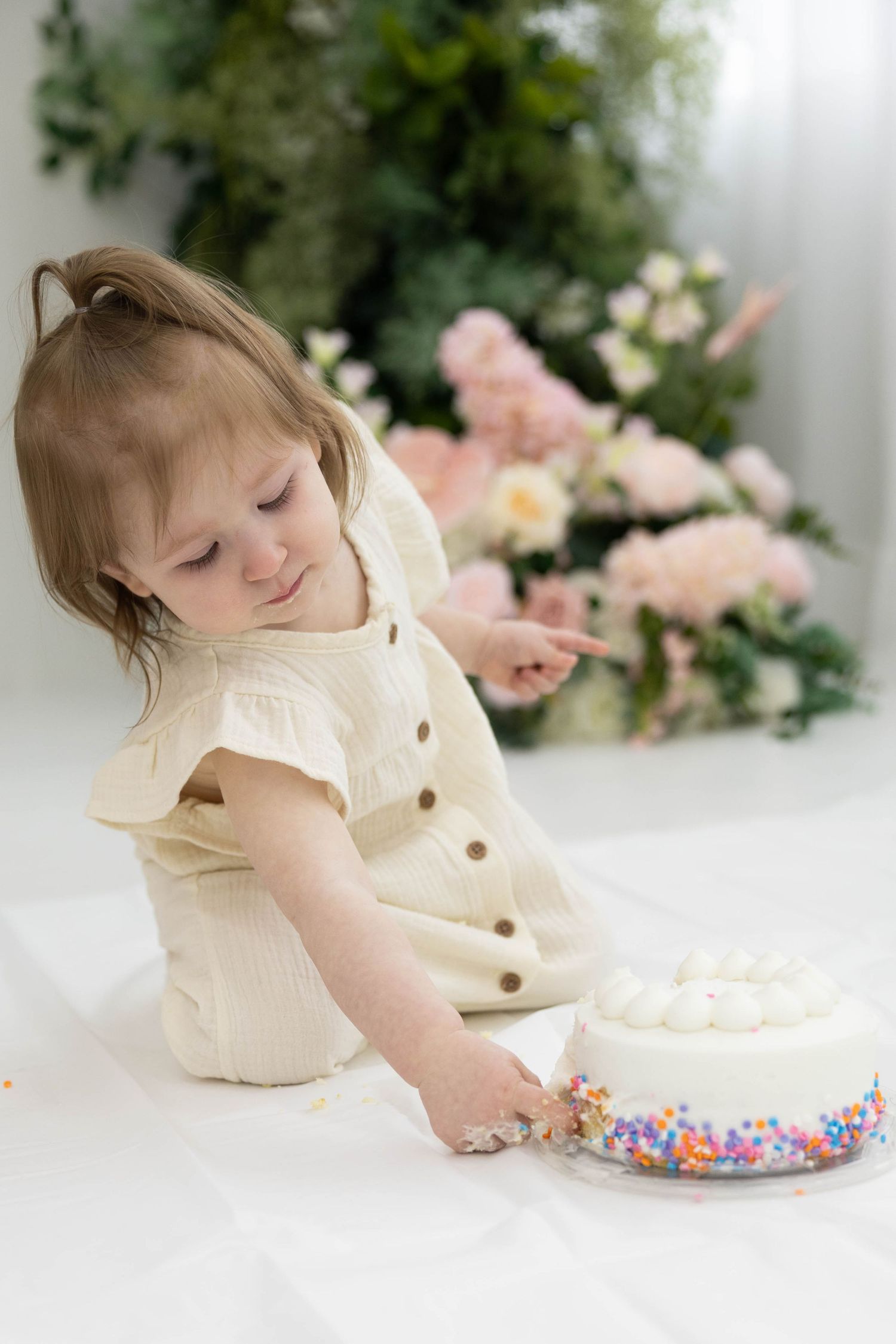 Toddler in cream outfit reaching for white cake with rainbow sprinkles against pink floral background.