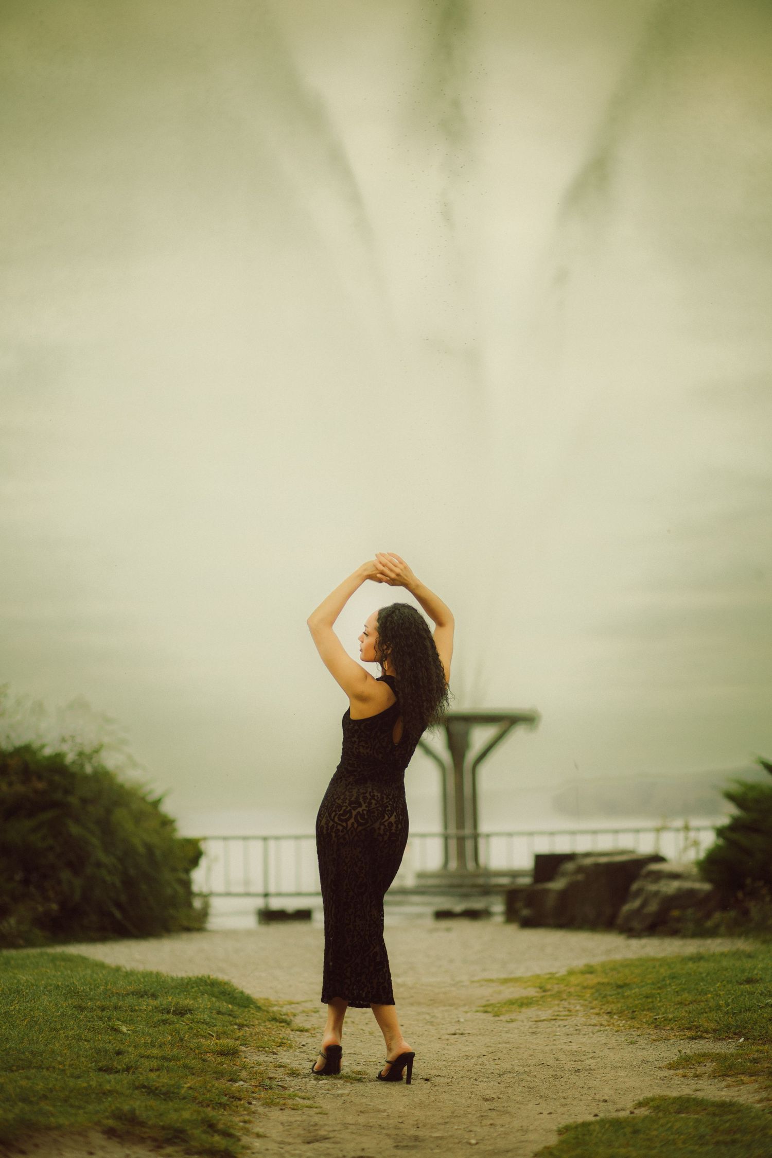 Woman in black dress poses gracefully with arms raised before a misty fountain in an outdoor park setting.