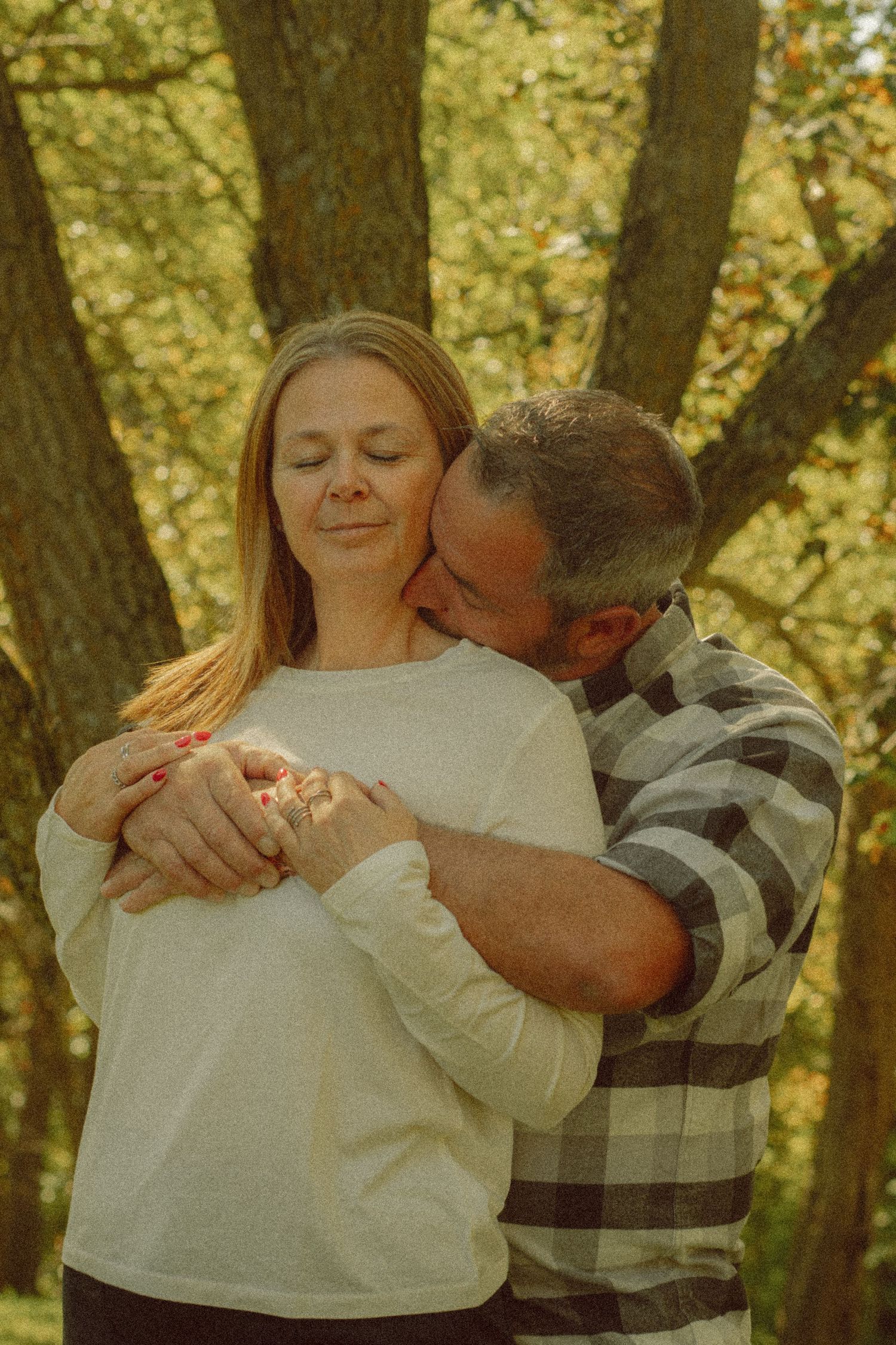 A couple embraces tenderly in an autumn park, man kissing woman's cheek while holding her hands.