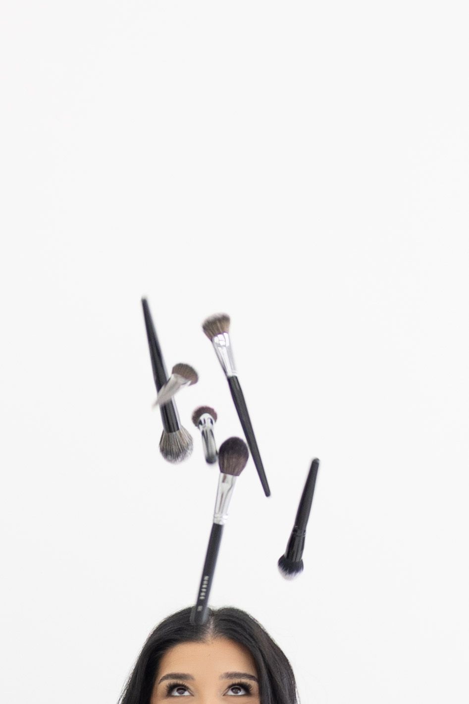 Woman looking up at falling makeup brushes on white background, beauty tools floating mid-air in creative cosmetics concept.