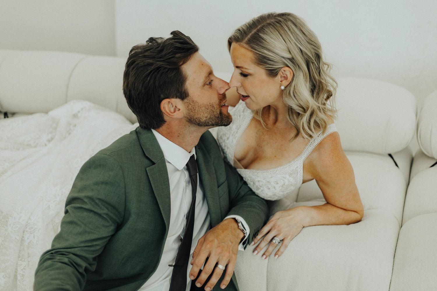 A romantic moment between a couple on a white couch, with one wearing a green blazer and the other in a white dress.